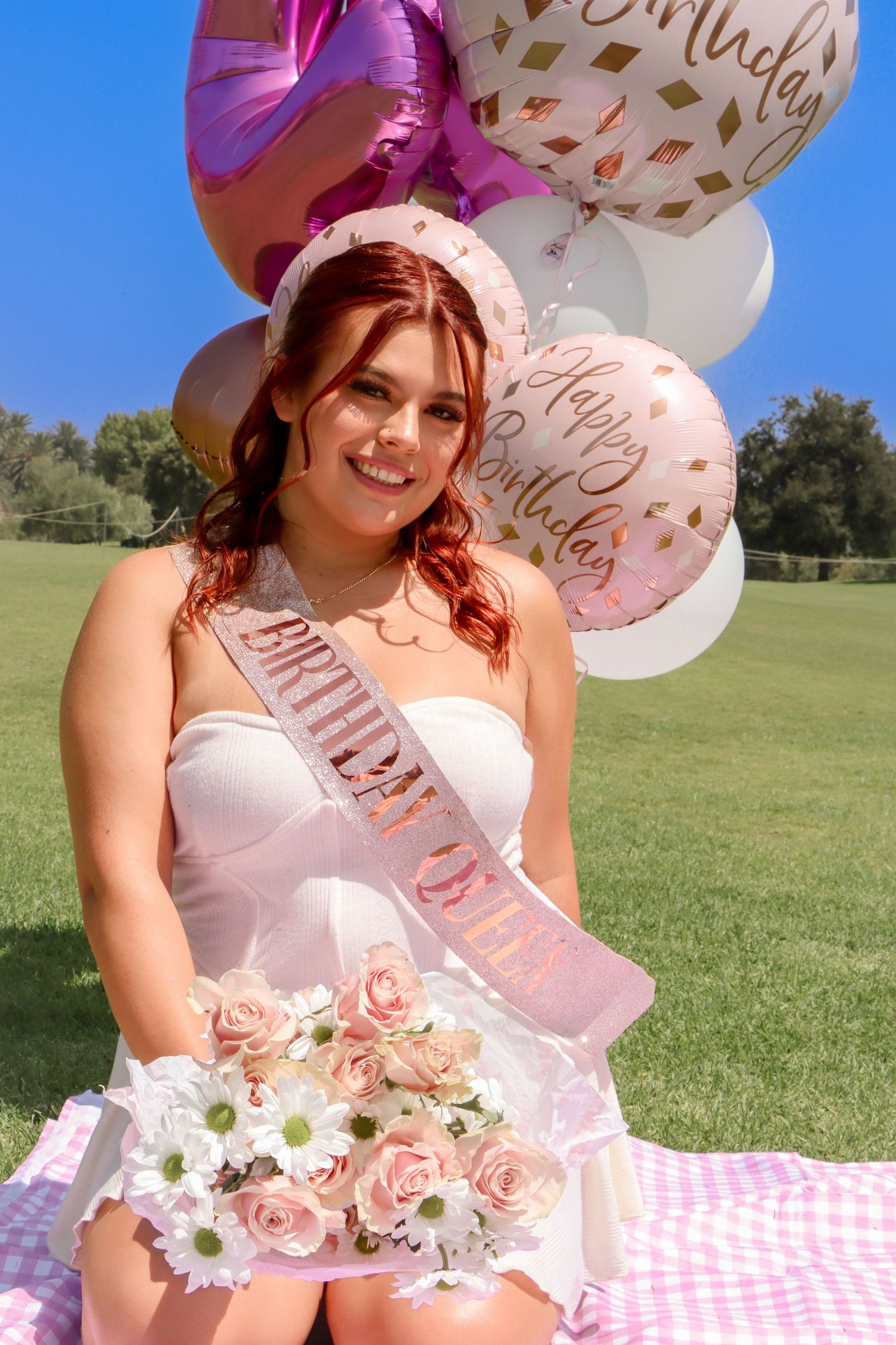 Birthday celebration outdoors with pink and white balloons and roses on checkered picnic blanket.