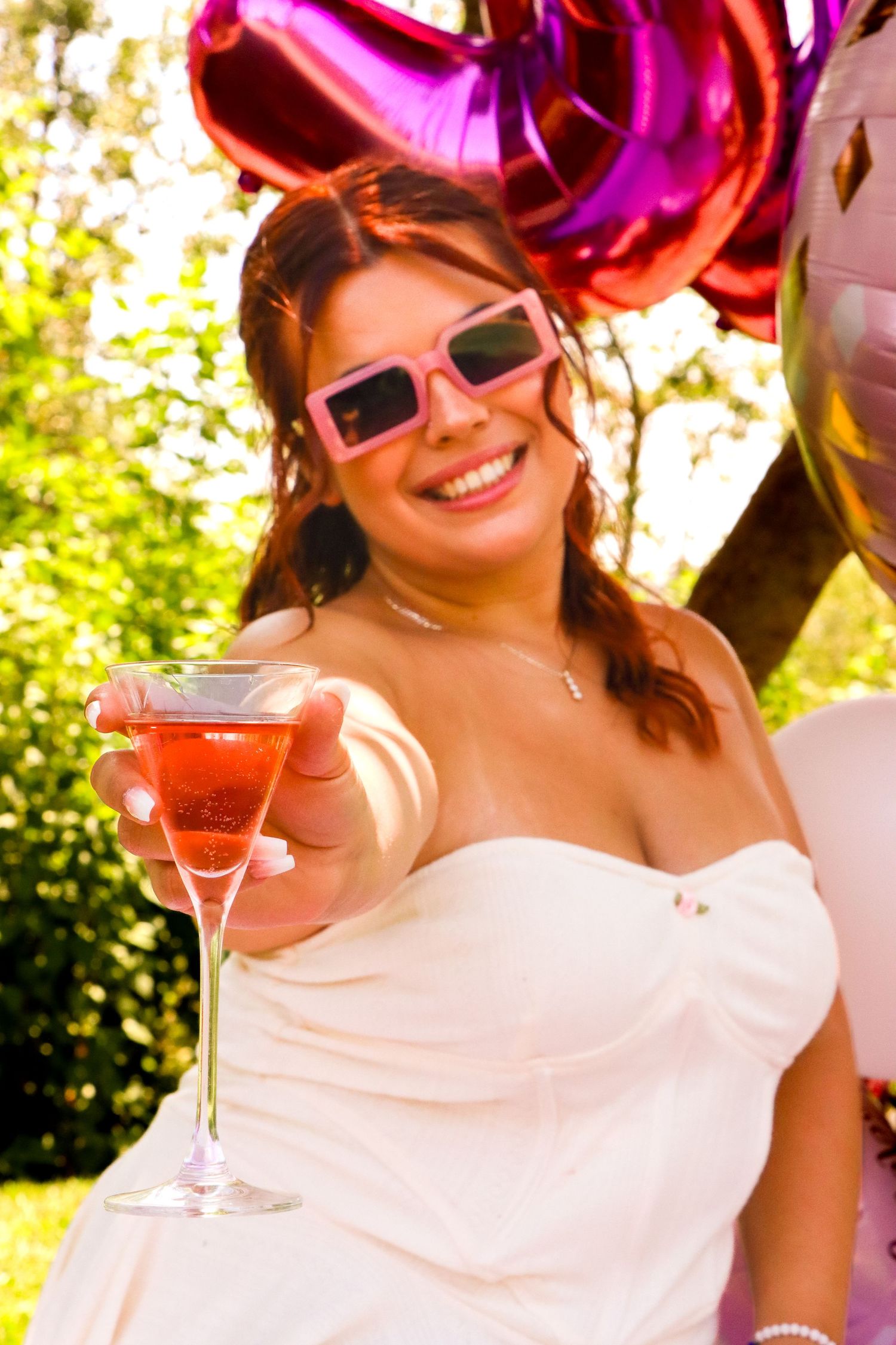 Person in white dress holding pink cocktail at outdoor celebration with balloons.