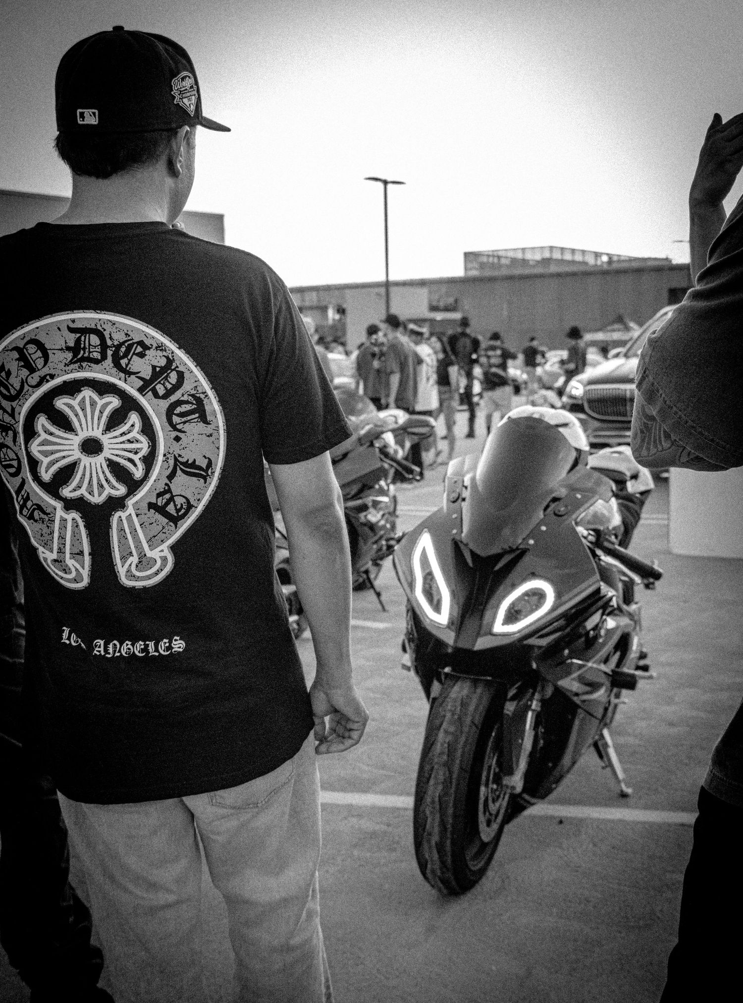 Man in Chrome Hearts LA tee stands beside a sport motorcycle in a black and white parking lot scene.