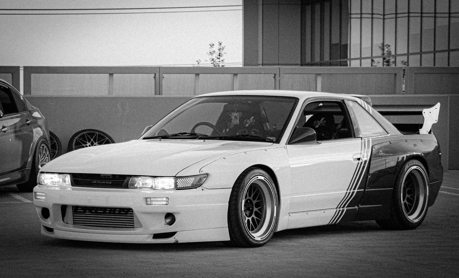 Modified white Nissan Silvia S13 with wide body kit, large rear spoiler, and deep dish wheels at a car meet.