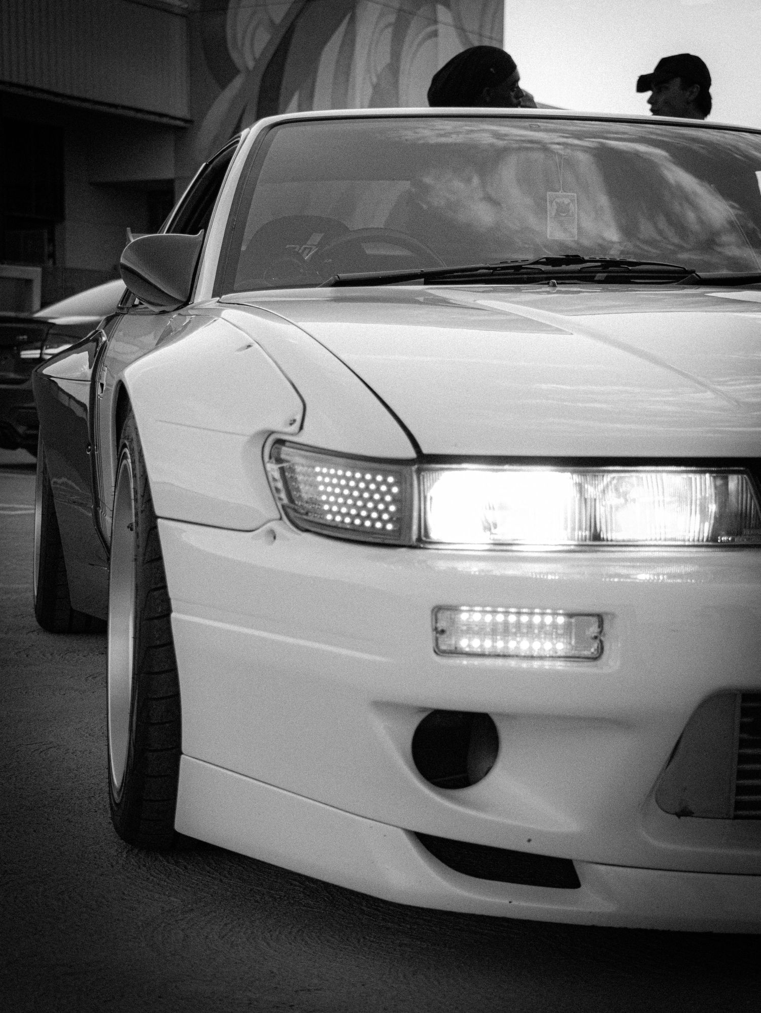 Black and white photo of a modified white Nissan Silvia S14 with LED headlights at a car meet.