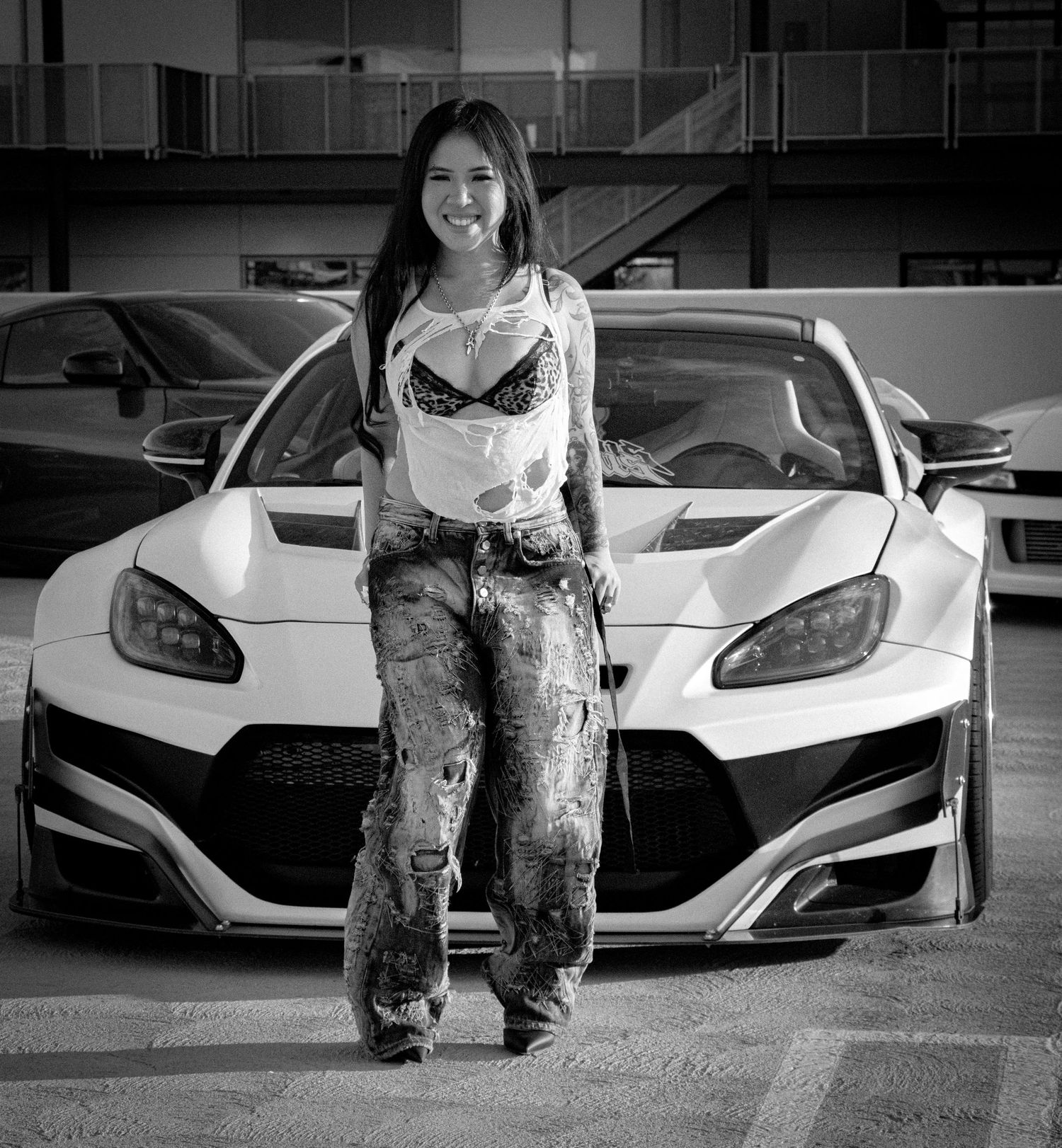 Young woman in ripped jeans poses in front of a white modified sports car in a parking lot, black and white photo.