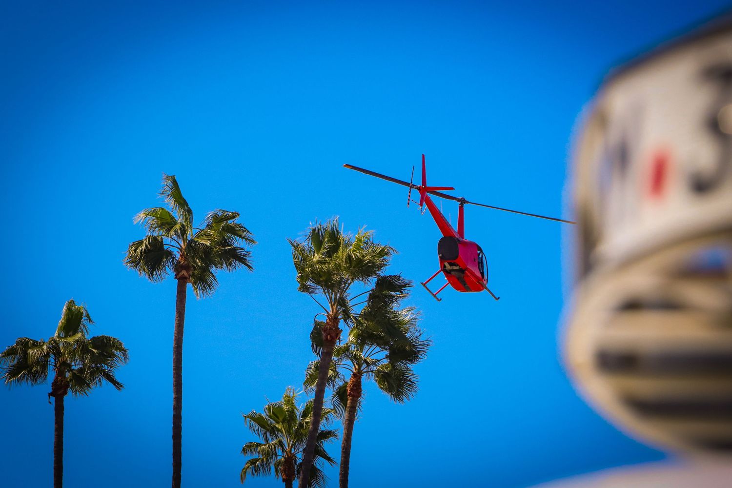 Red helicopter flying against bright blue sky with palm trees below.