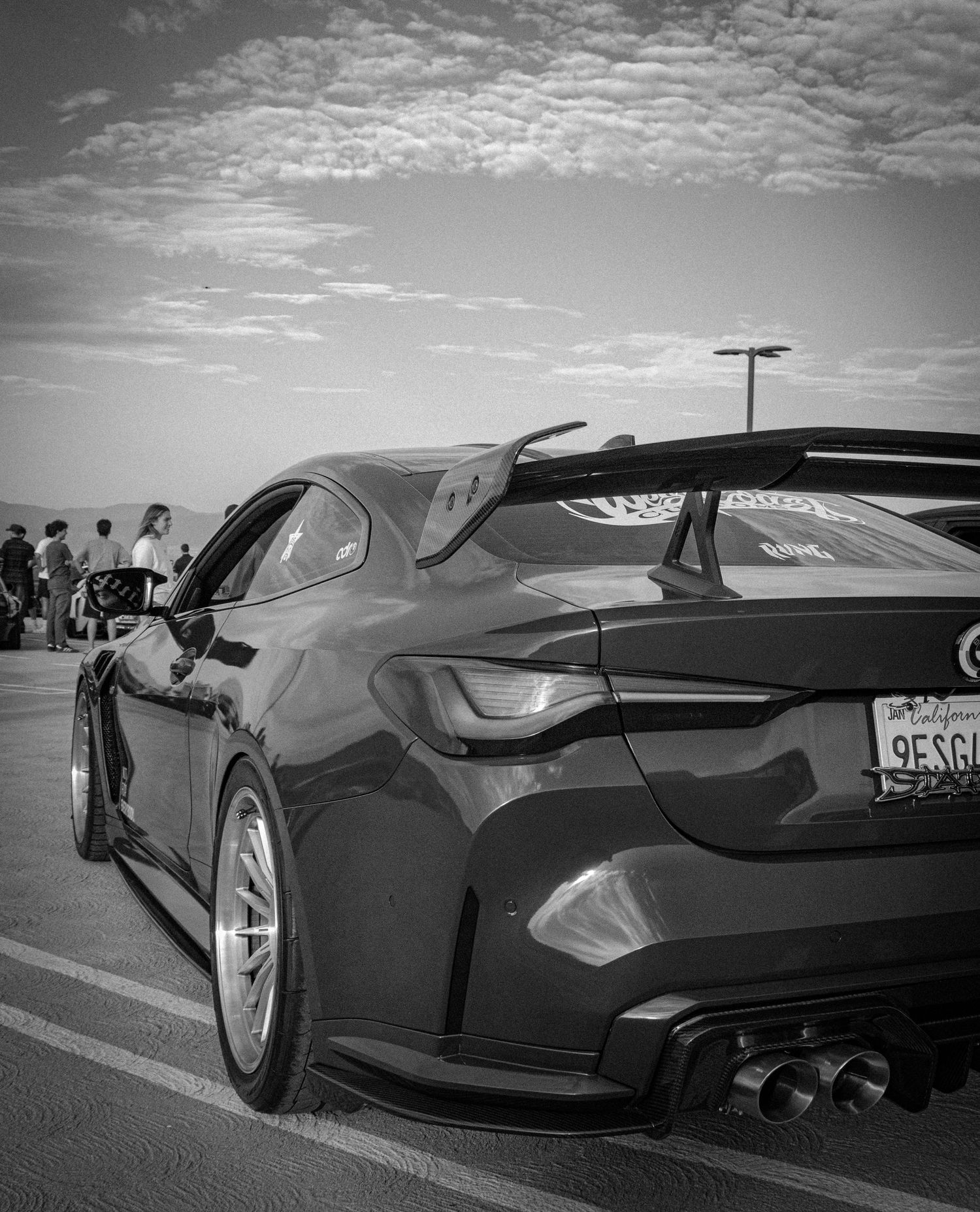 Black and white photo of a modified sports car with large rear spoiler and quad exhaust tips in a parking lot.