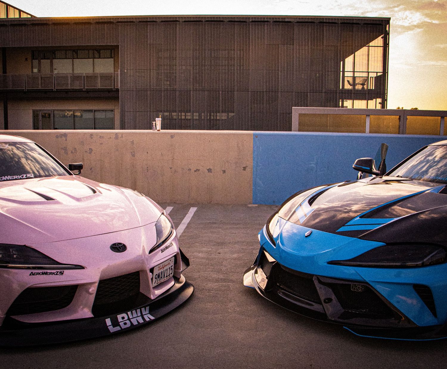Two modified sports cars, a pink Toyota Supra and blue Corvette C8, parked side by side at a rooftop location.