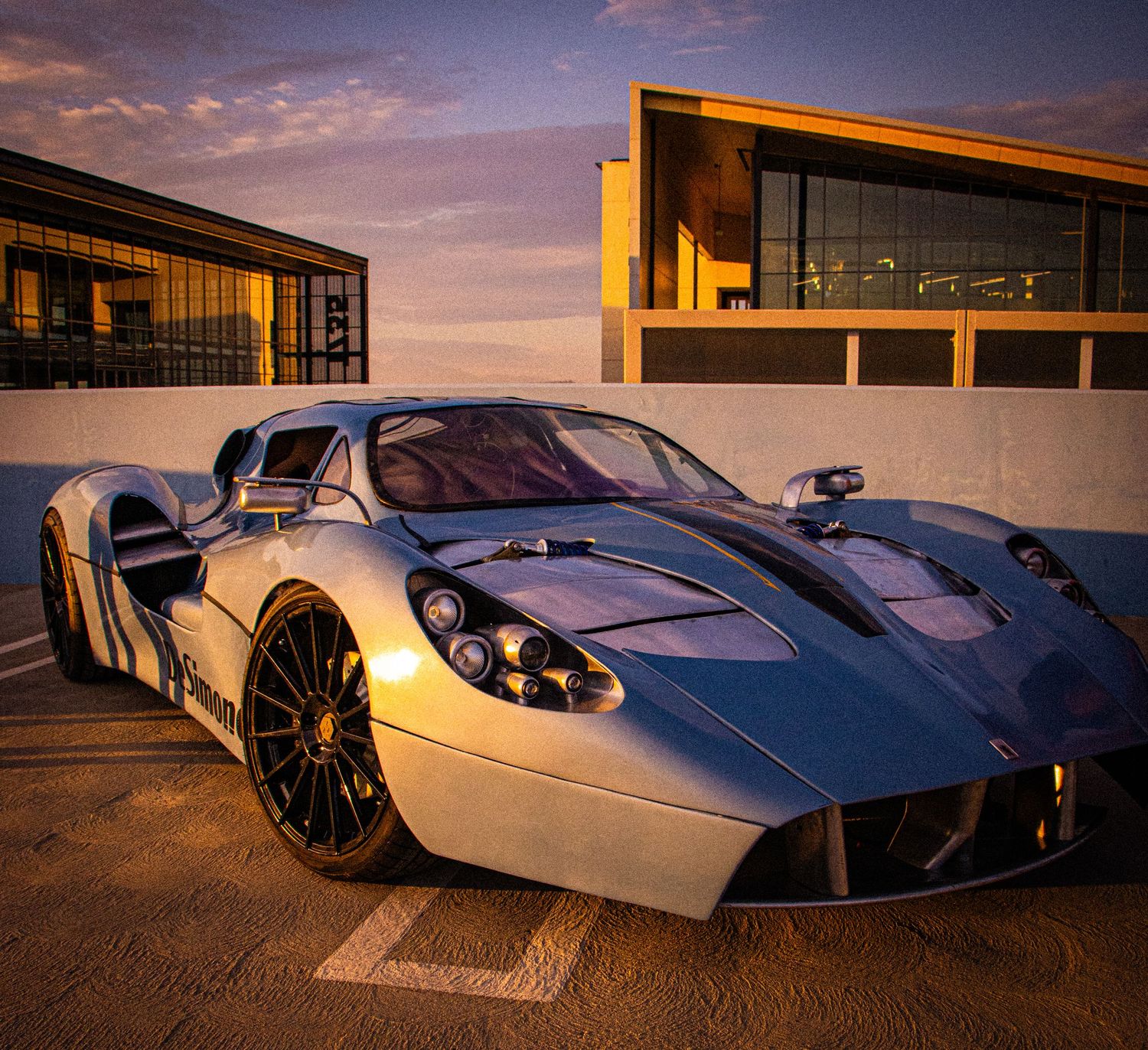 Silver supercar parked at sunset with dramatic purple sky and modern building backdrop.
