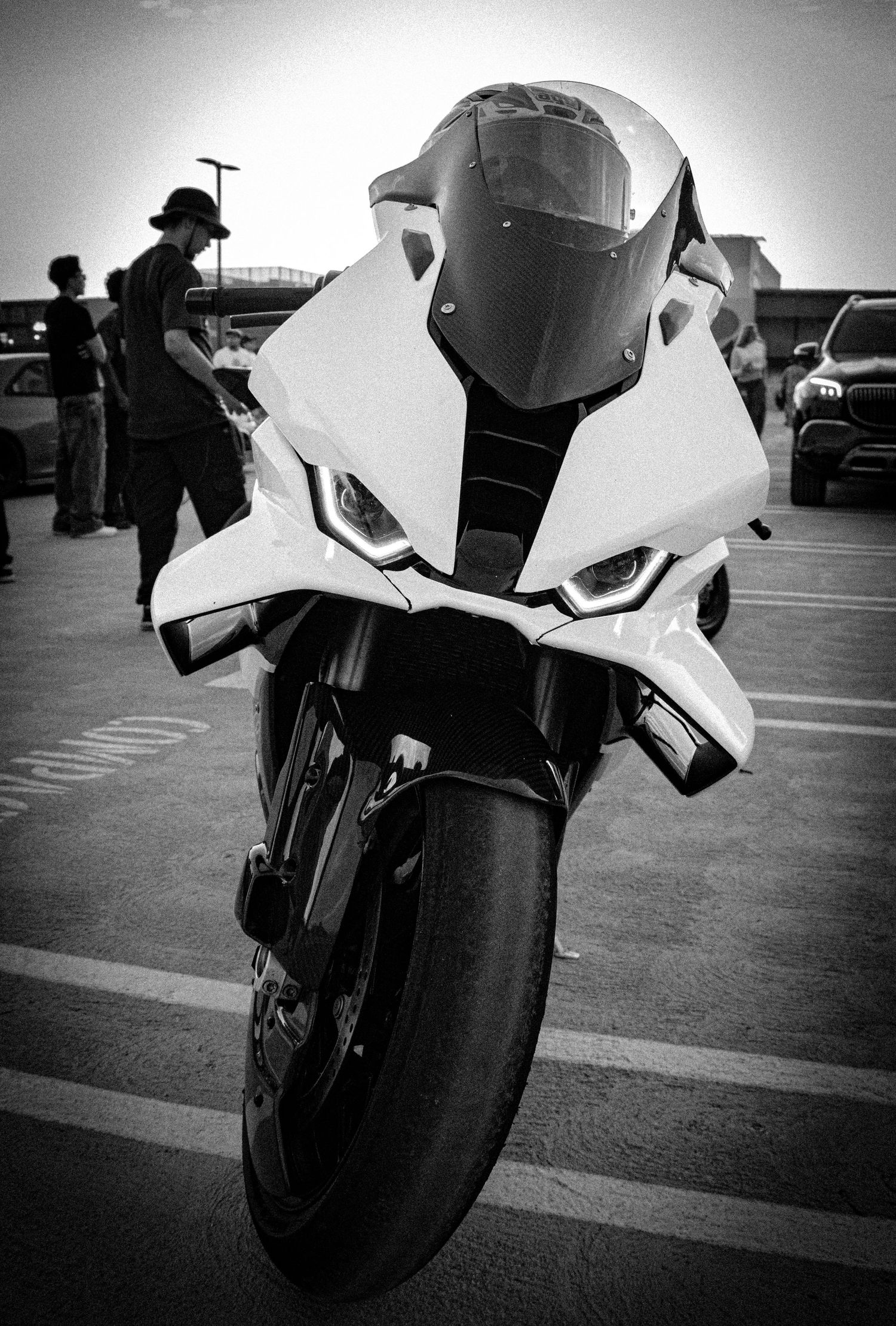 Black and white front view of a sport motorcycle parked in a lot, with people and cars visible in the background.