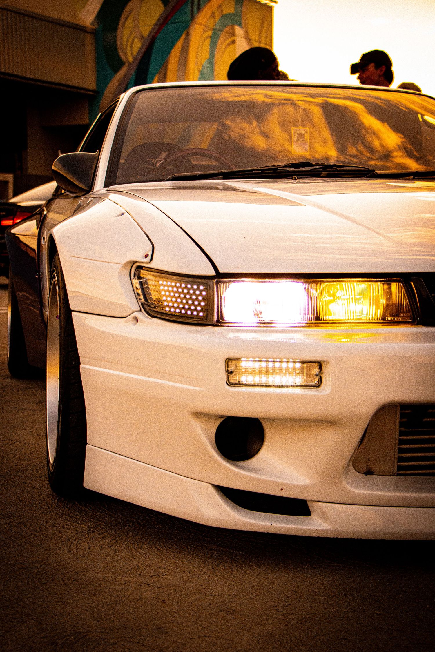 White modified Nissan Silvia S13 with glowing amber headlights and lowered stance at a car meet.