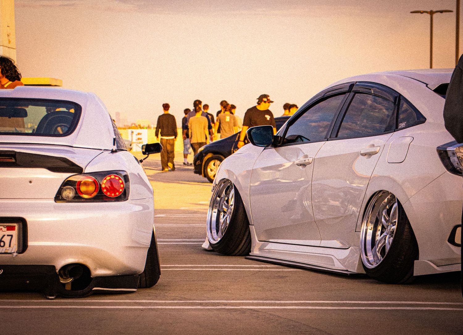 Two lowered white sports cars parked at a car meet during golden hour with spectators in the background.