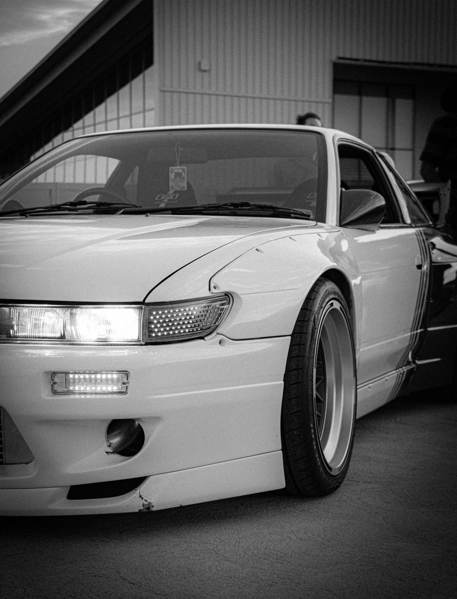 Black and white photo of a modified Nissan S13 Silvia with wide body kit and deep dish wheels at a car show.