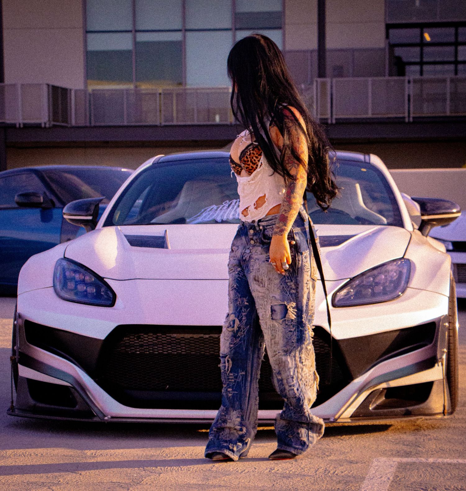 Woman in ripped jeans posing in front of a white widebody sports car in a parking lot at dusk.