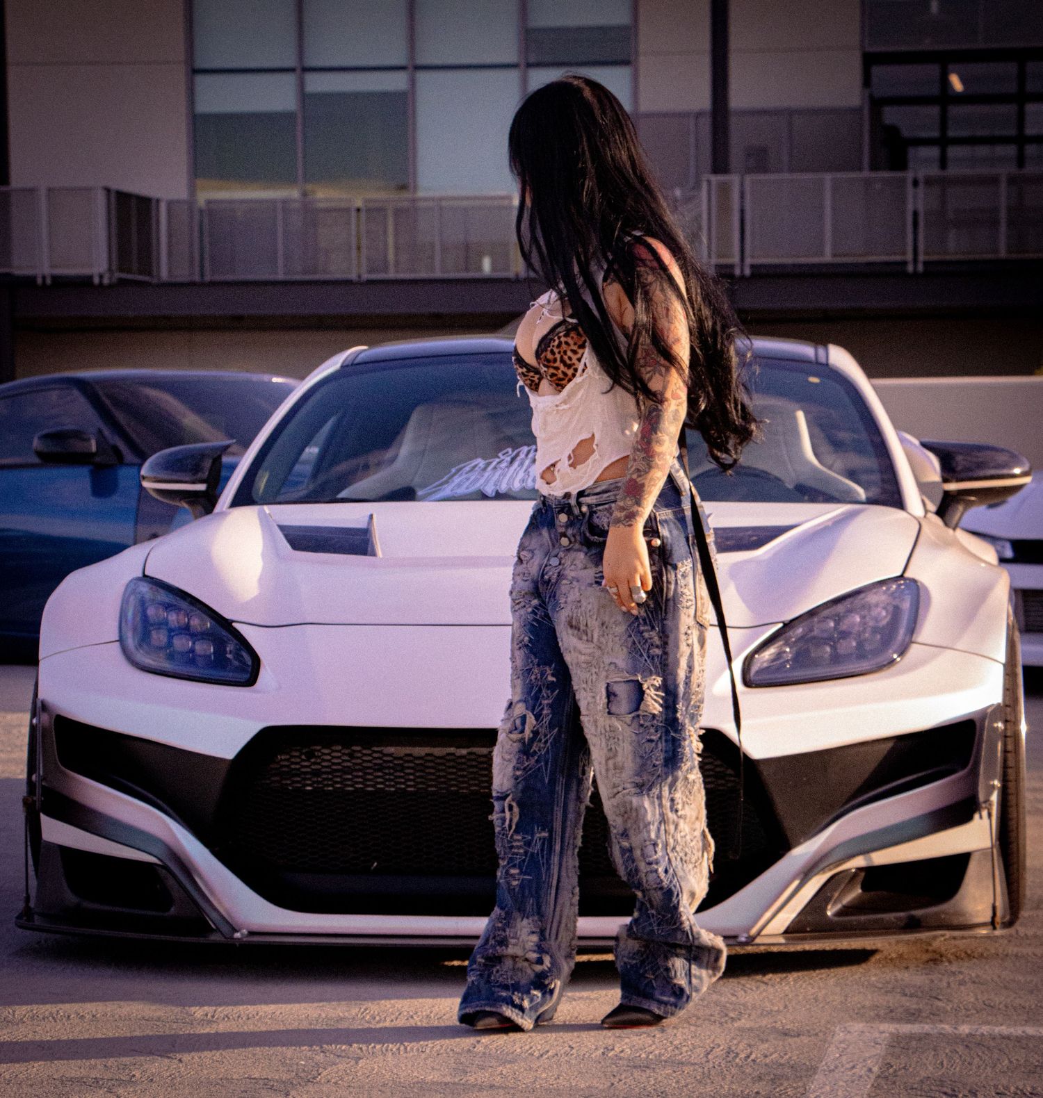 Woman in ripped jeans posing in front of a white widebody sports car in a parking lot.