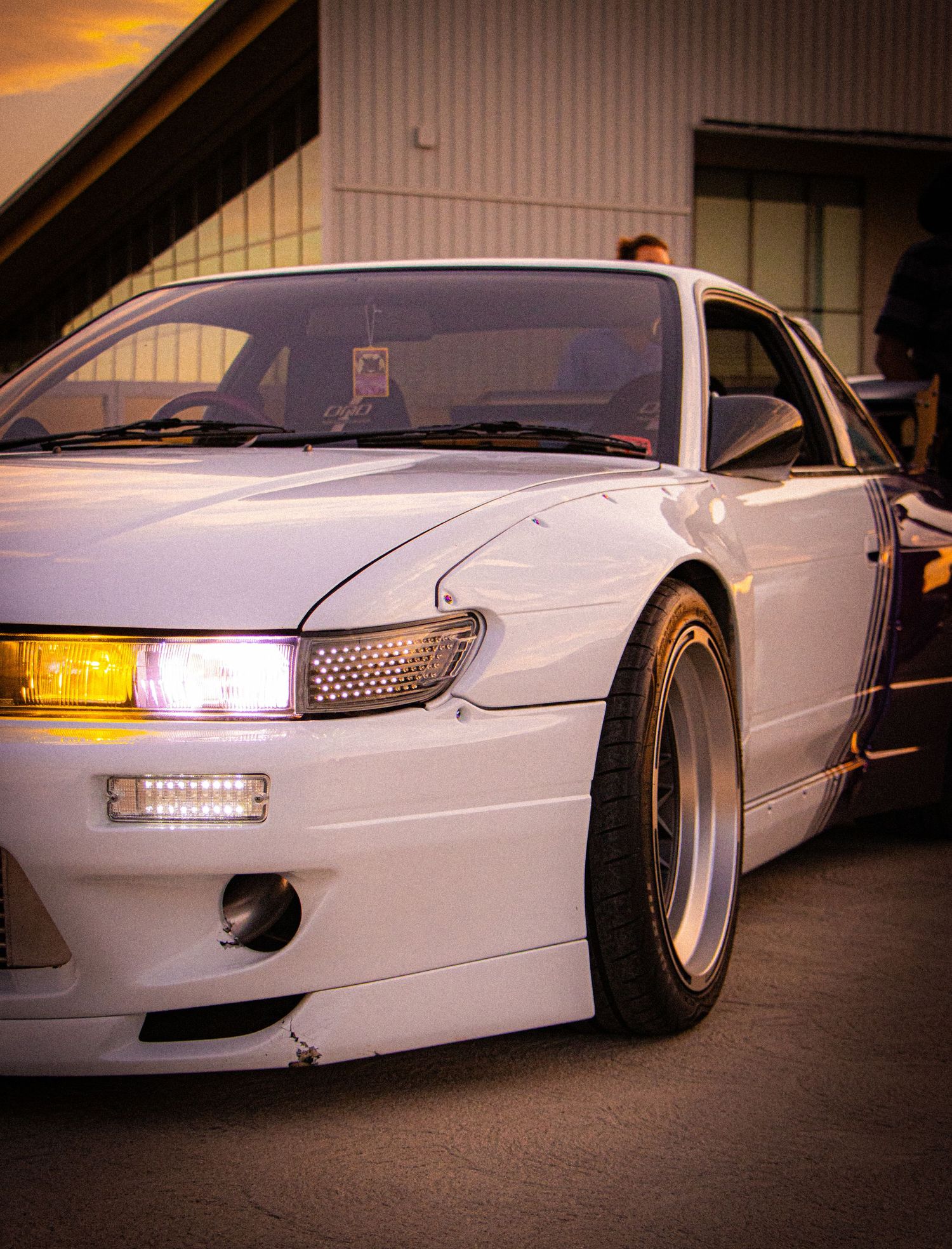 White Nissan S13 silvia with yellow headlights and wide body kit parked at sunset during a car meet.