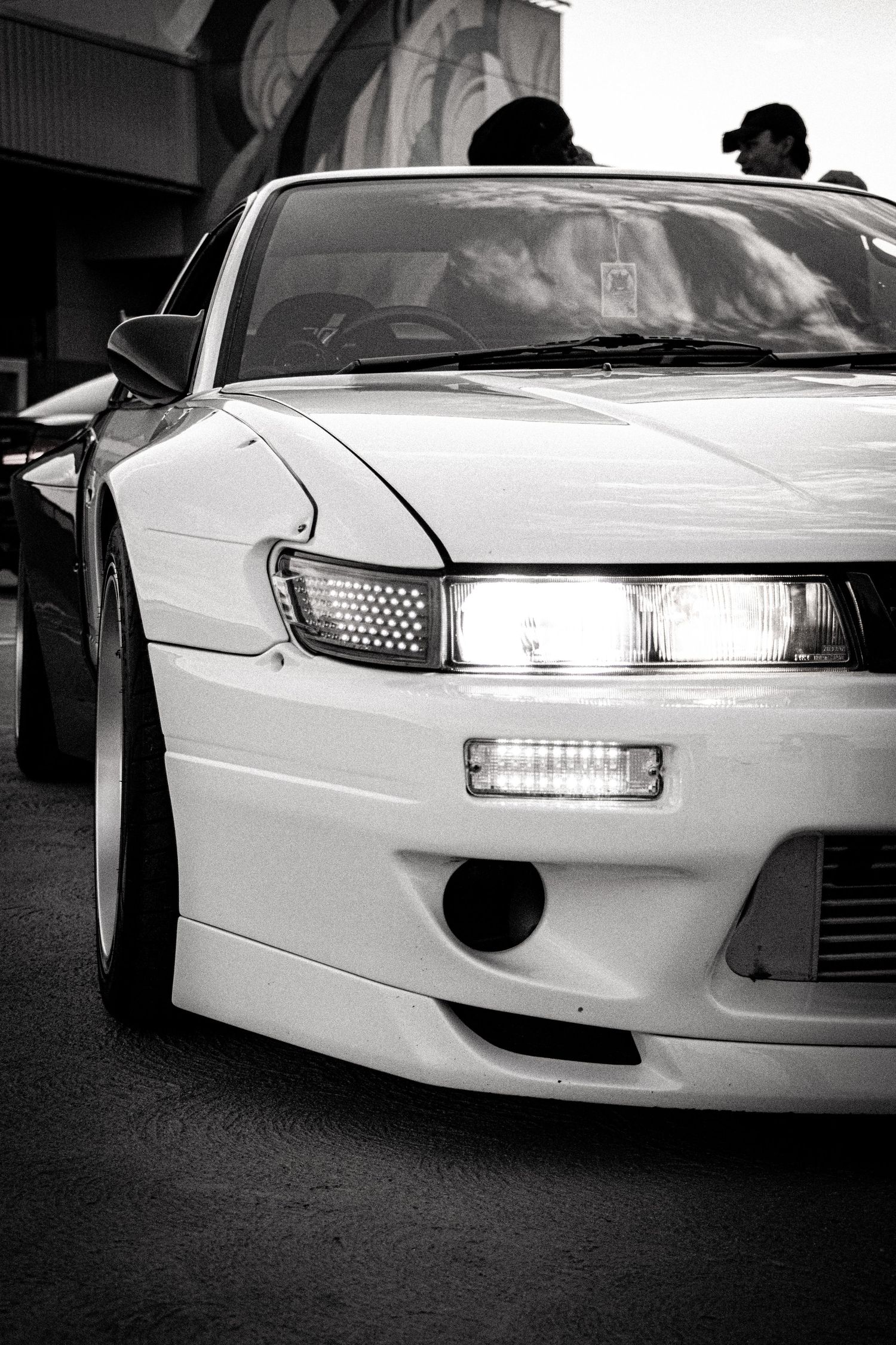Black and white close-up of a modified white Nissan S13 Silvia with aftermarket bumper and stanced low profile stance.