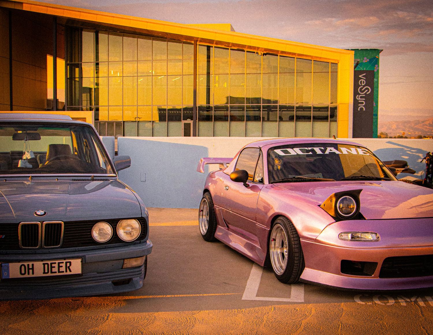 A blue BMW E30 and pink Mazda Miata with Octane branding parked outside a modern building at sunset.