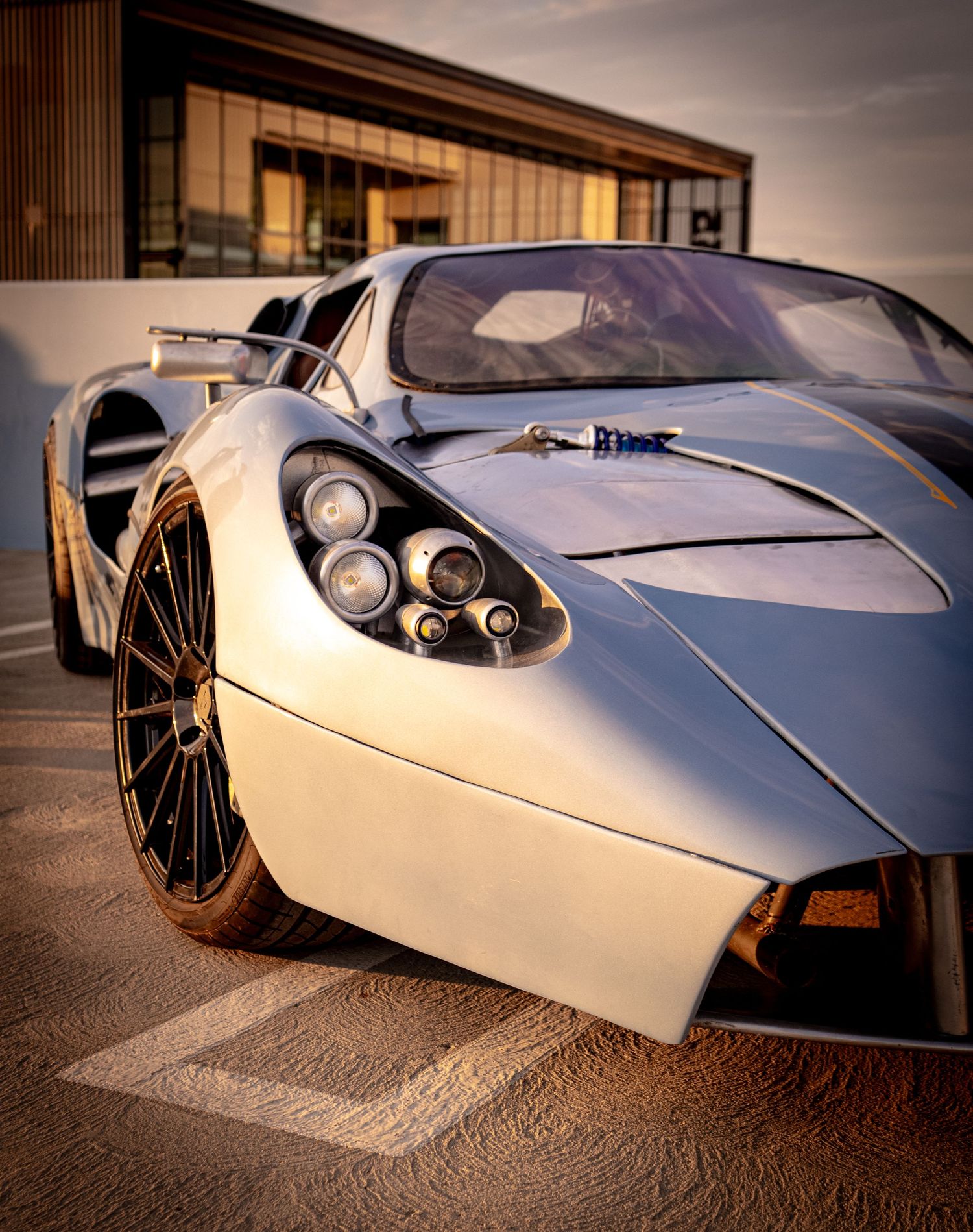 A sleek white supercar with aggressive styling and large alloy wheels parked outside a modern building at golden hour.