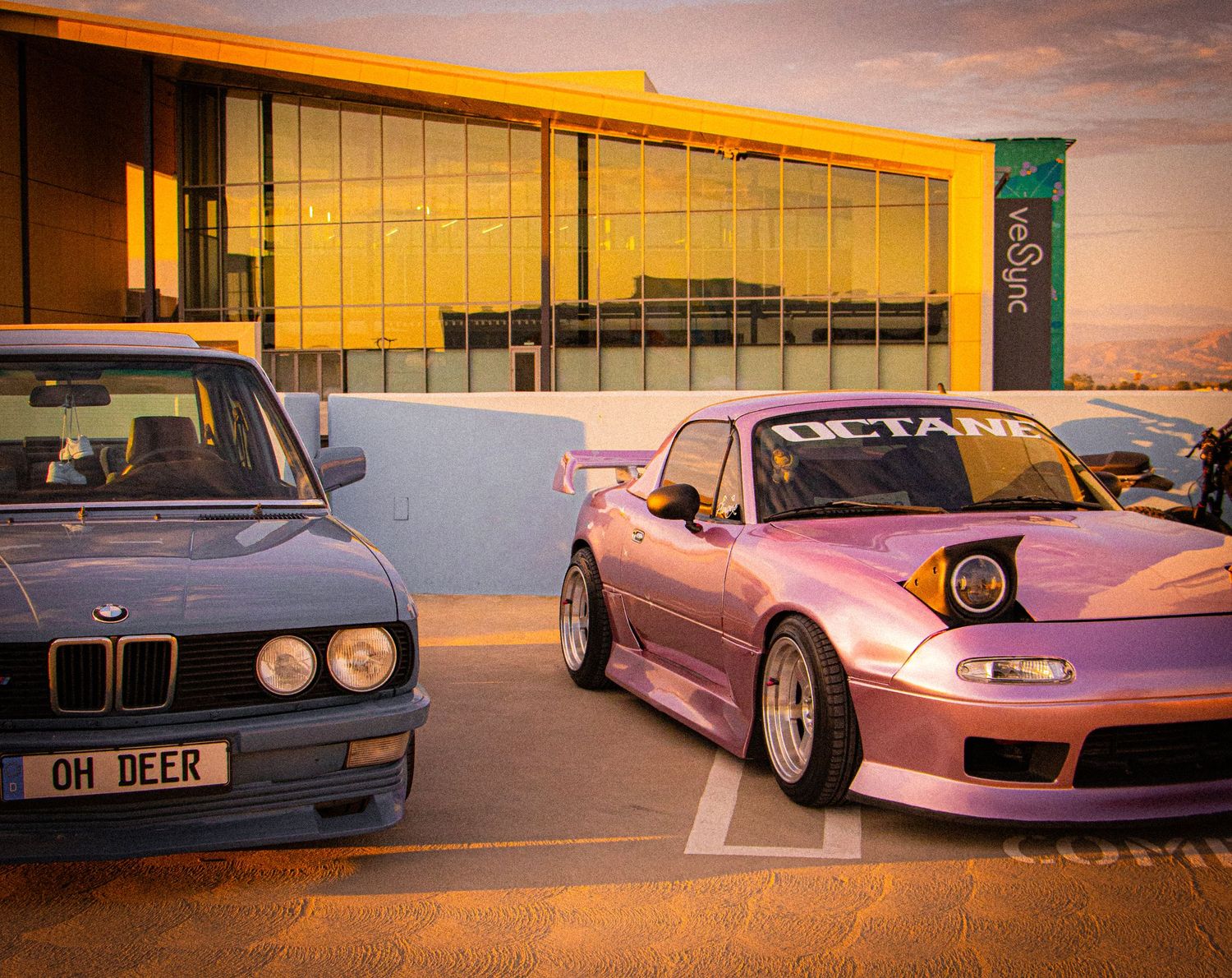 A blue BMW E28 and pink widebody Mazda MX-5 parked at sunset outside a modern building.