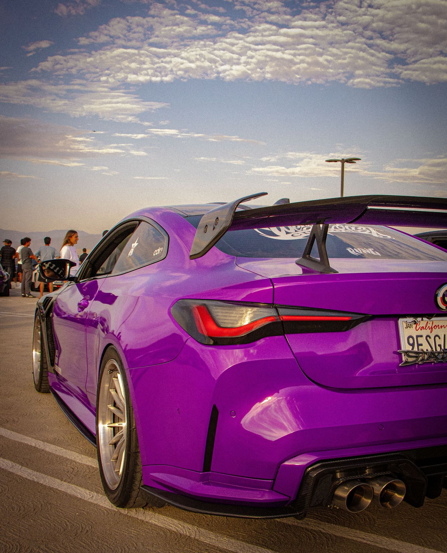 Vibrant purple BMW M4 with large rear spoiler and quad exhaust tips parked at a car meet under a cloudy sky.