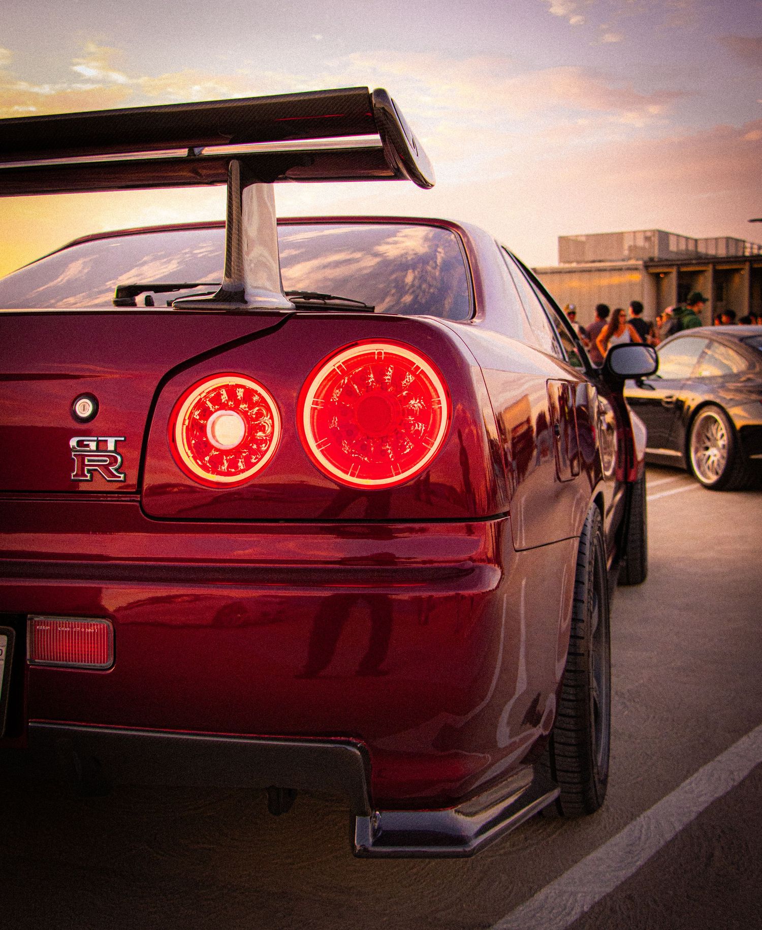 Red Nissan Skyline GT-R R34 rear view with glowing taillights at sunset in a parking lot.