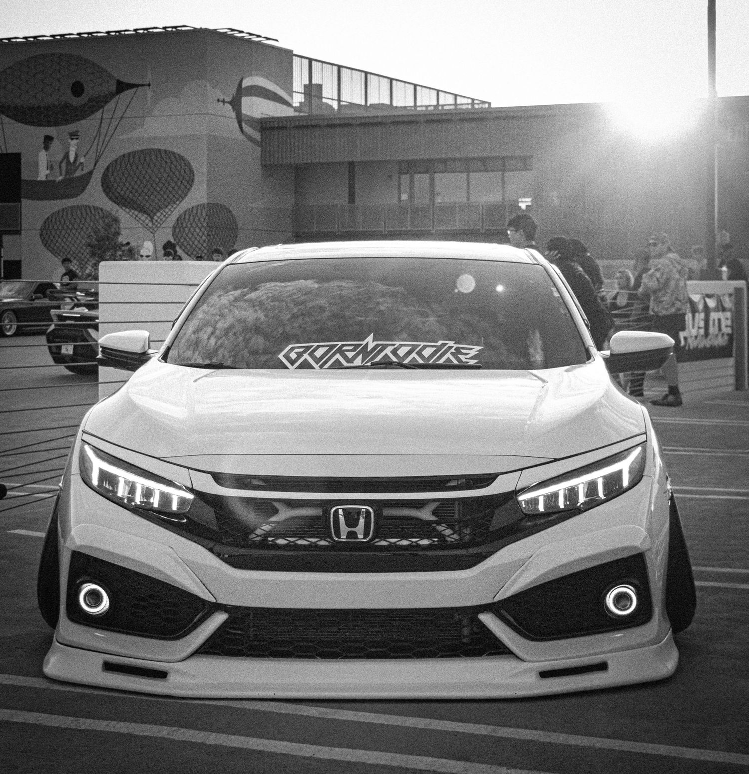 Black and white photo of a modified Honda Civic with custom body kit and LED headlights at a car show.