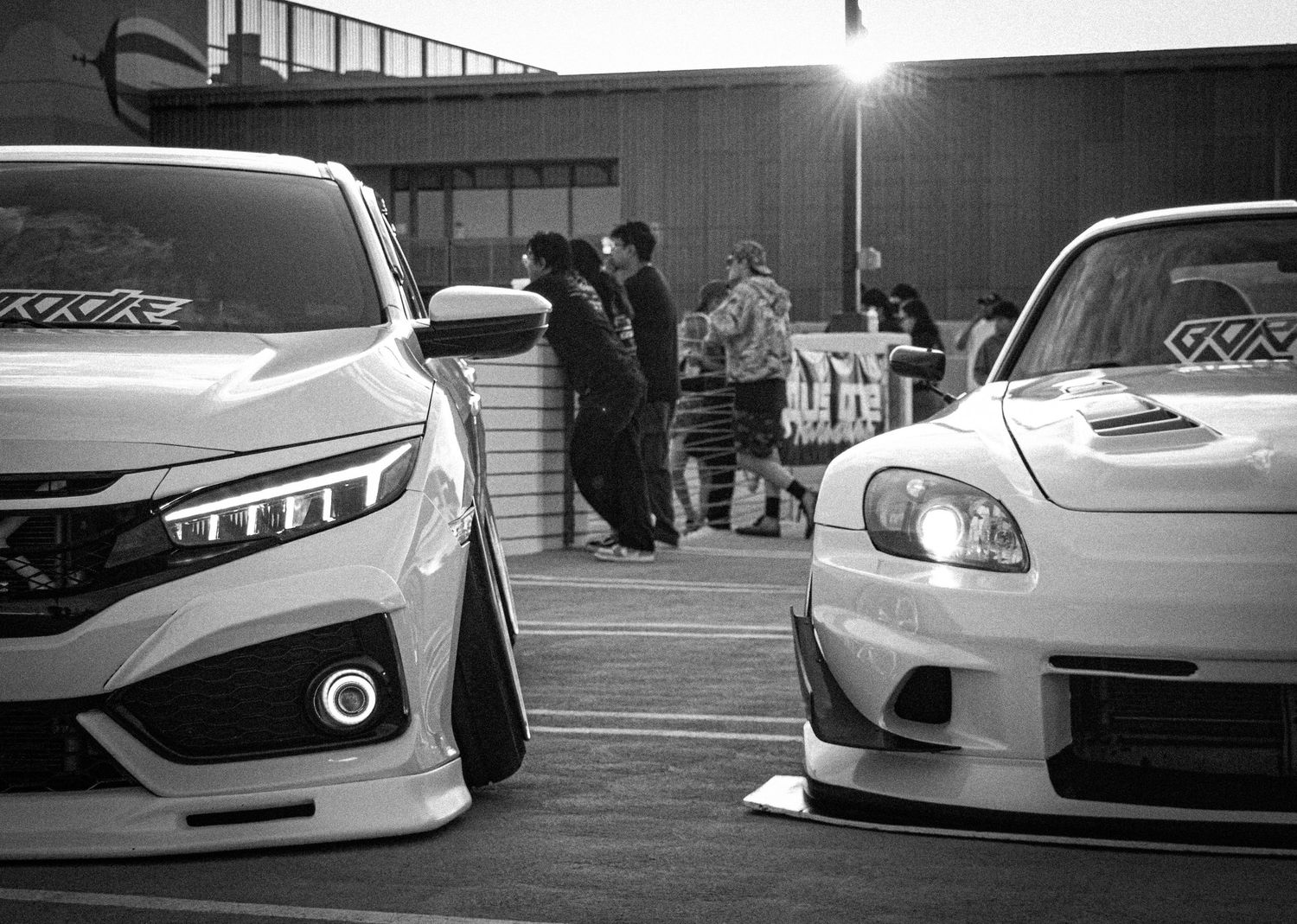 Black and white photo of two modified Honda cars at a car meet with people gathered in the background.