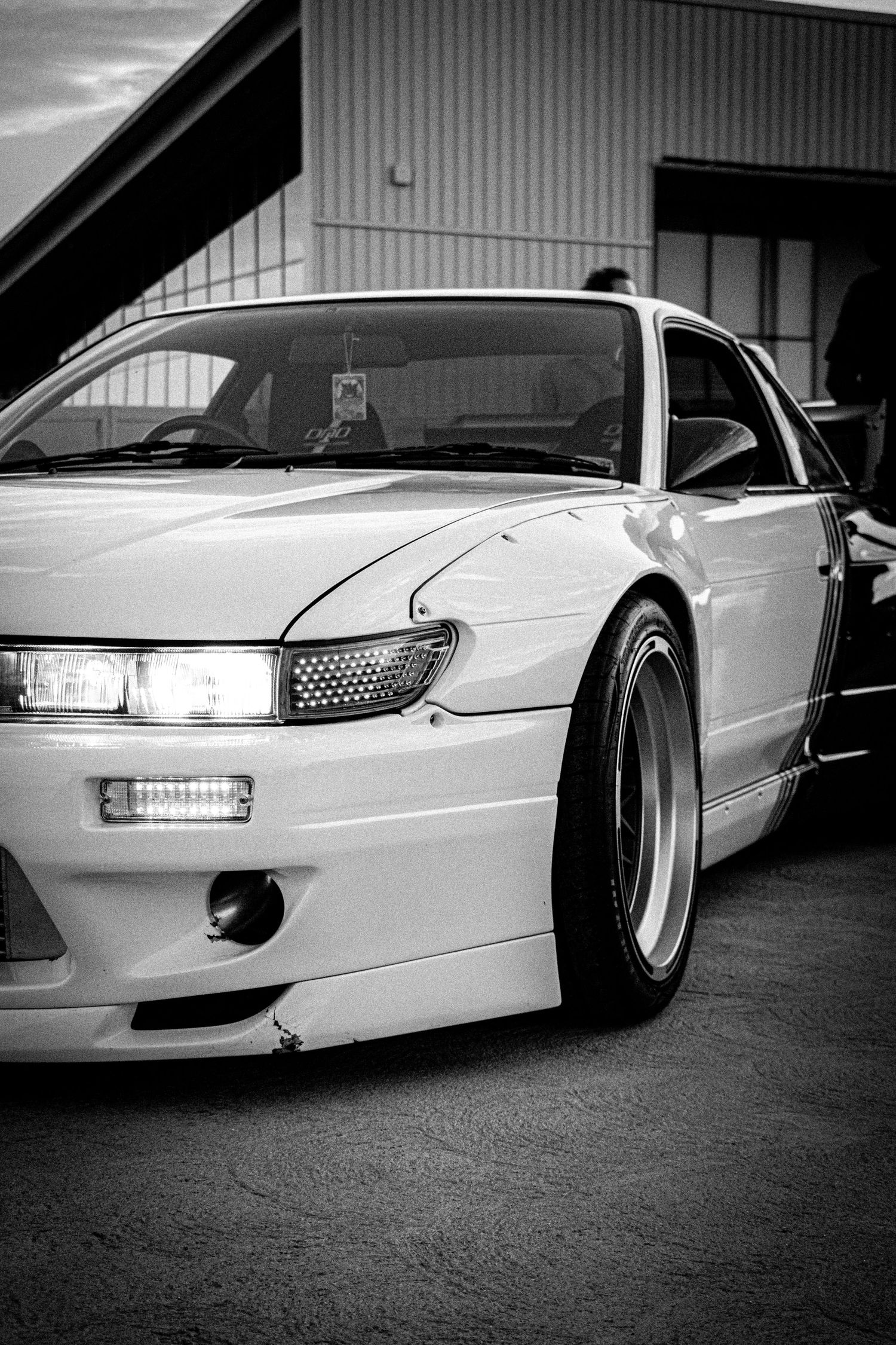 Black and white photo of a modified Nissan S13 Silvia with wide body kit and deep dish wheels parked outside.