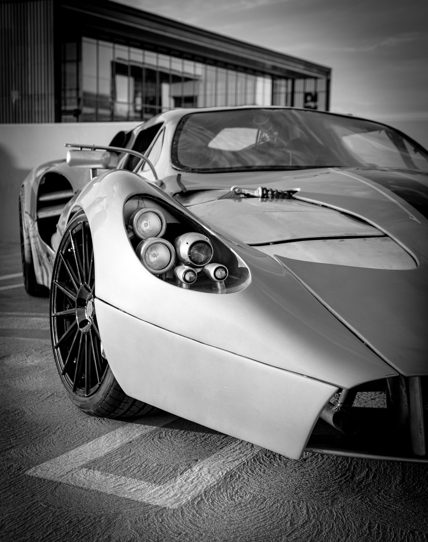 Black and white photo of a sleek supercar with quad exhausts parked in a lot with a modern building backdrop.