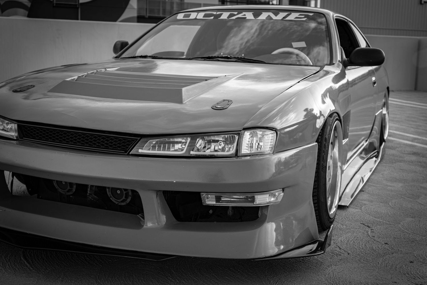Black and white photo of a modified Nissan S14 240SX with wide body kit and custom front bumper at a car show.