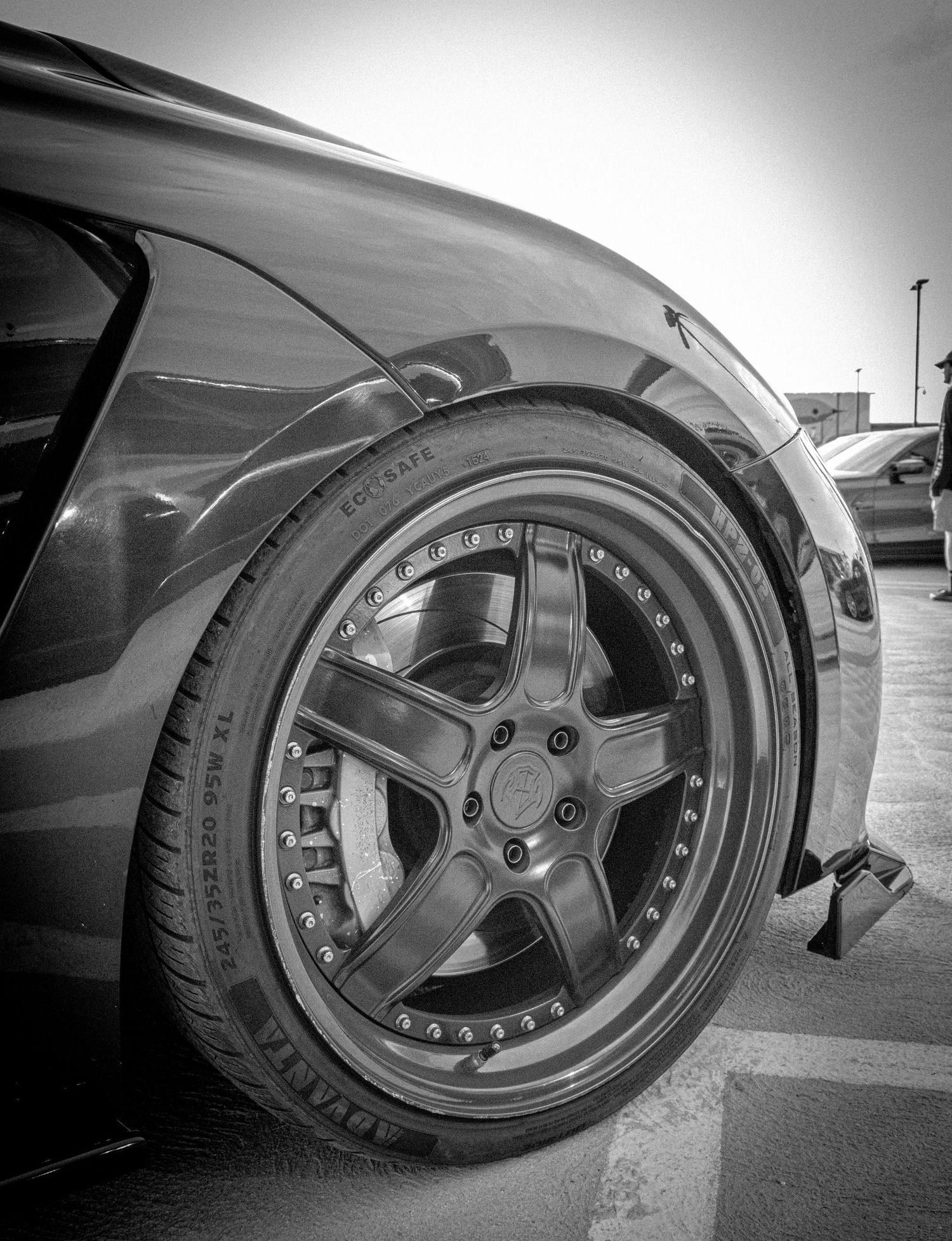 Black and white close-up of a custom multi-spoke alloy wheel on a lowered sports car in a parking lot.