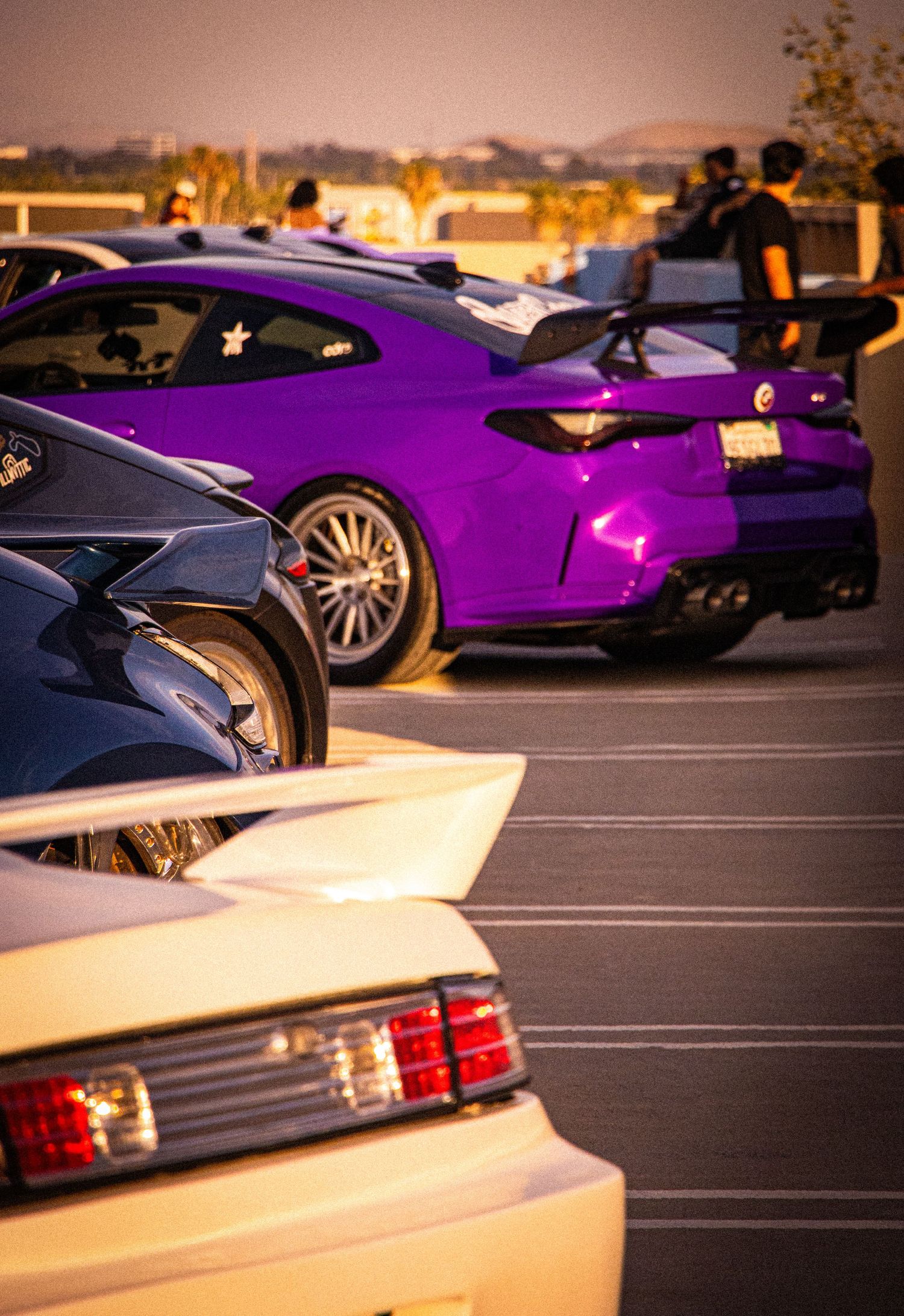 Purple sports car with large rear spoiler parked at a car meet, surrounded by other modified vehicles at dusk.