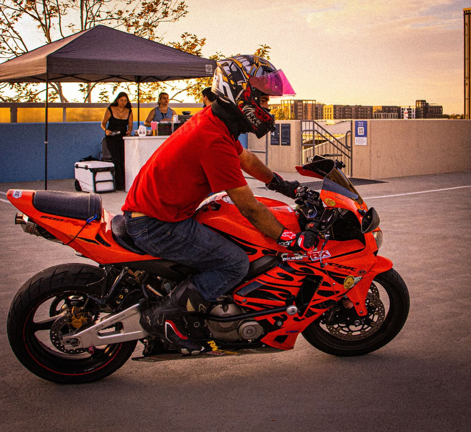 A motorcyclist in a red shirt and helmet rides a red sport motorcycle at an outdoor event during sunset.