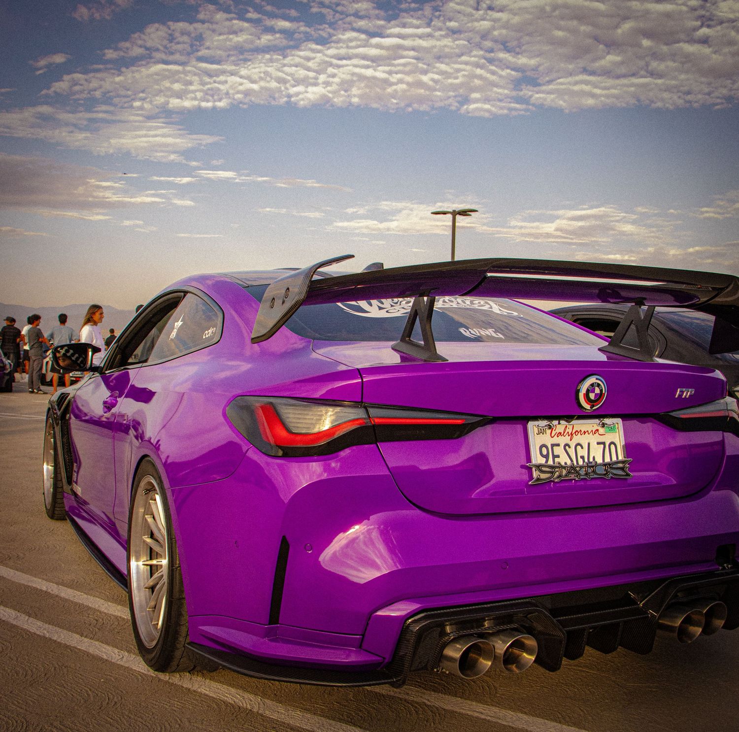Purple BMW M4 with wide body kit, large rear wing, and quad exhaust tips parked at a car show under blue skies.