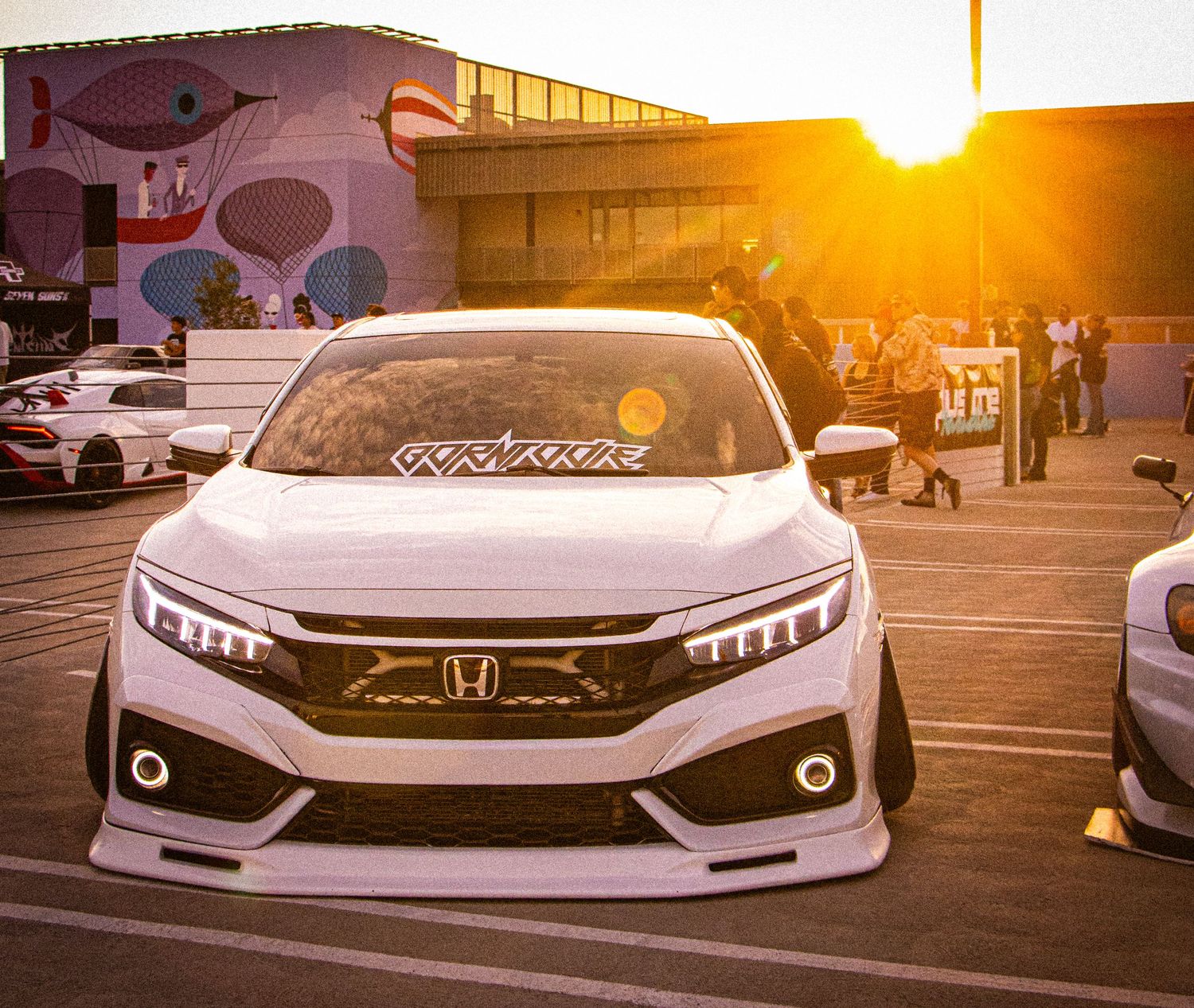 Modified white Honda Civic with custom body kit at a car show during golden hour sunset.