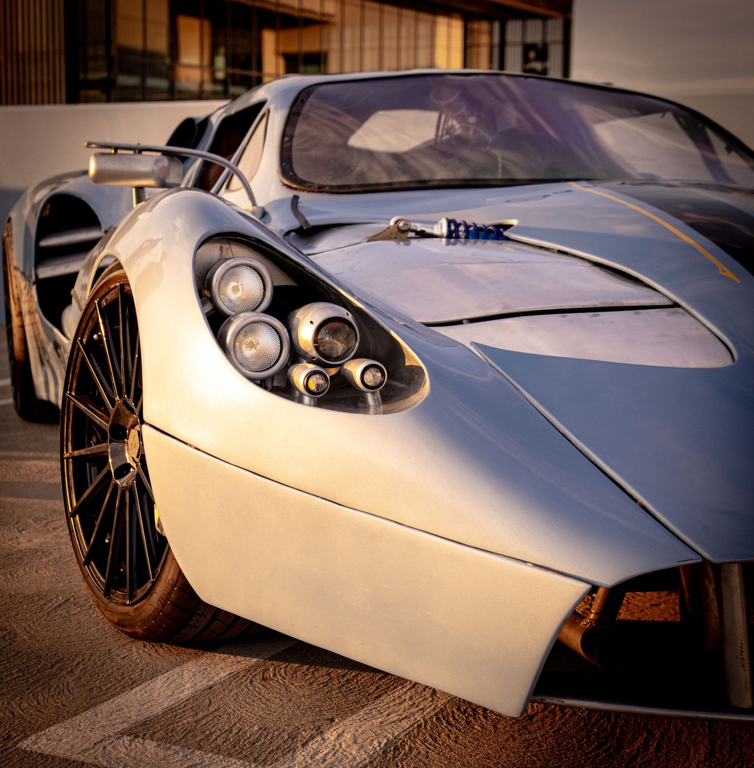 A sleek white Pagani supercar with quad headlights and gold accents parked outdoors in warm sunlight.