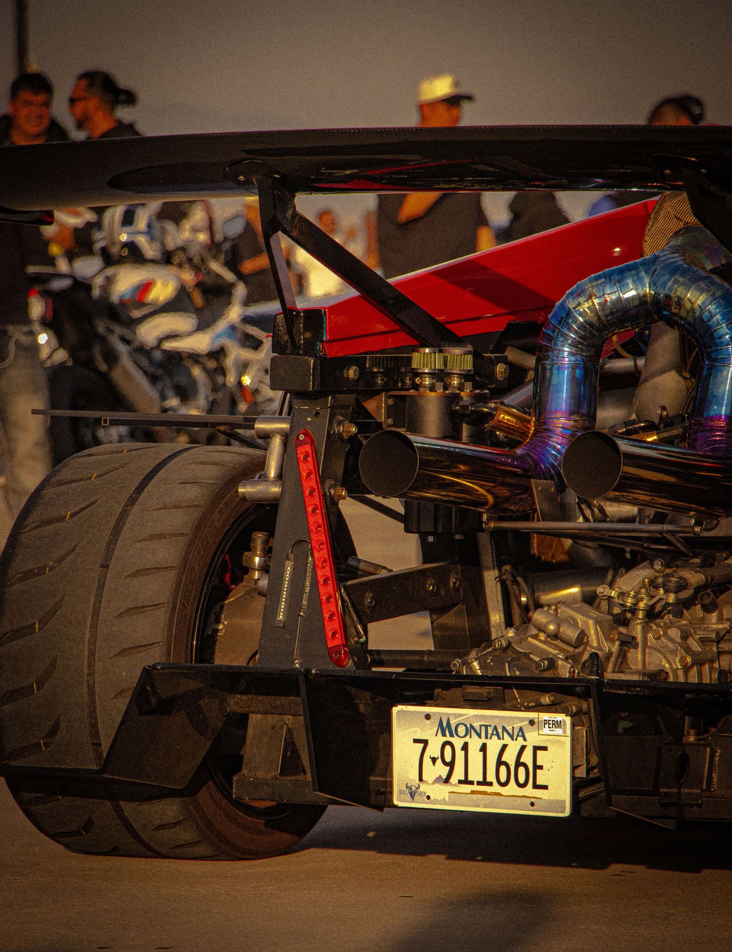 Custom Montana-plated drag racing car with colorful exhaust pipes and wide rear tires at a nighttime motorsport event.