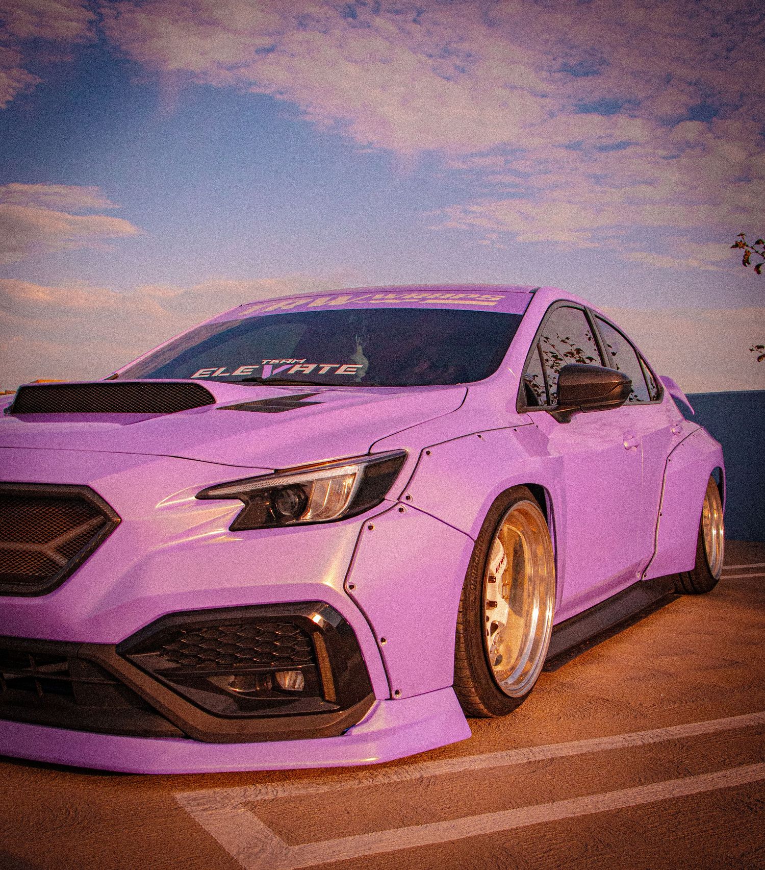 Pink widebody Subaru WRX STI with gold wheels and aggressive body kit, lowered stance, parked near ocean at sunset.