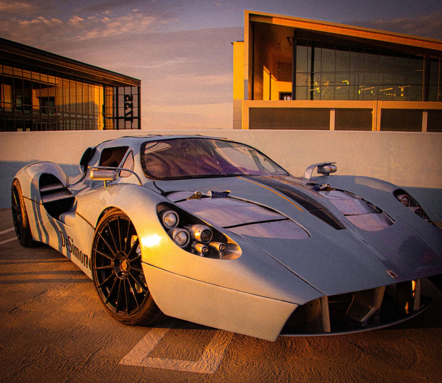 A sleek silver supercar parked at golden hour with modern buildings in the background.