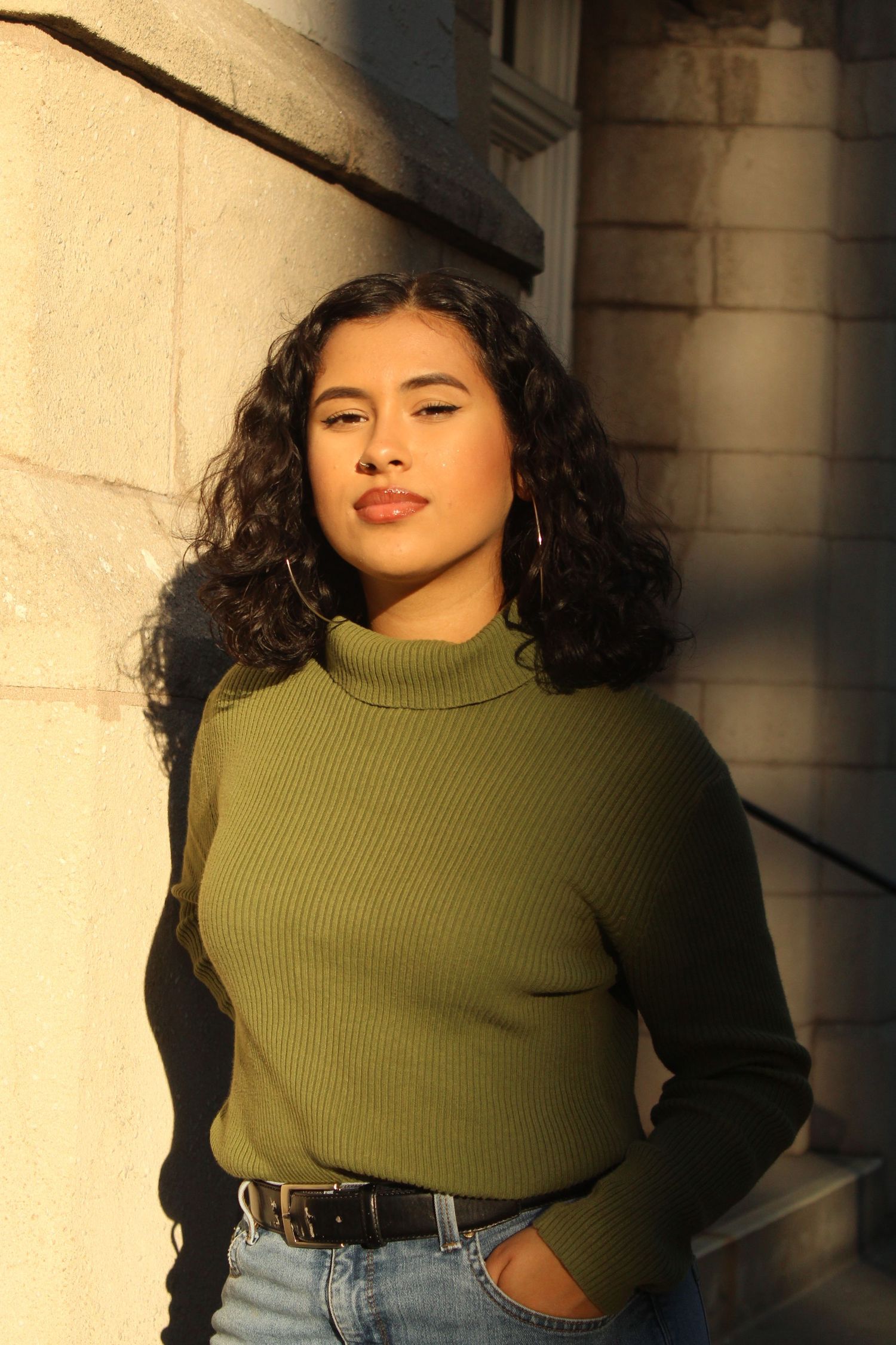 Woman in olive green turtleneck sweater and jeans poses against beige wall in warm sunlight.