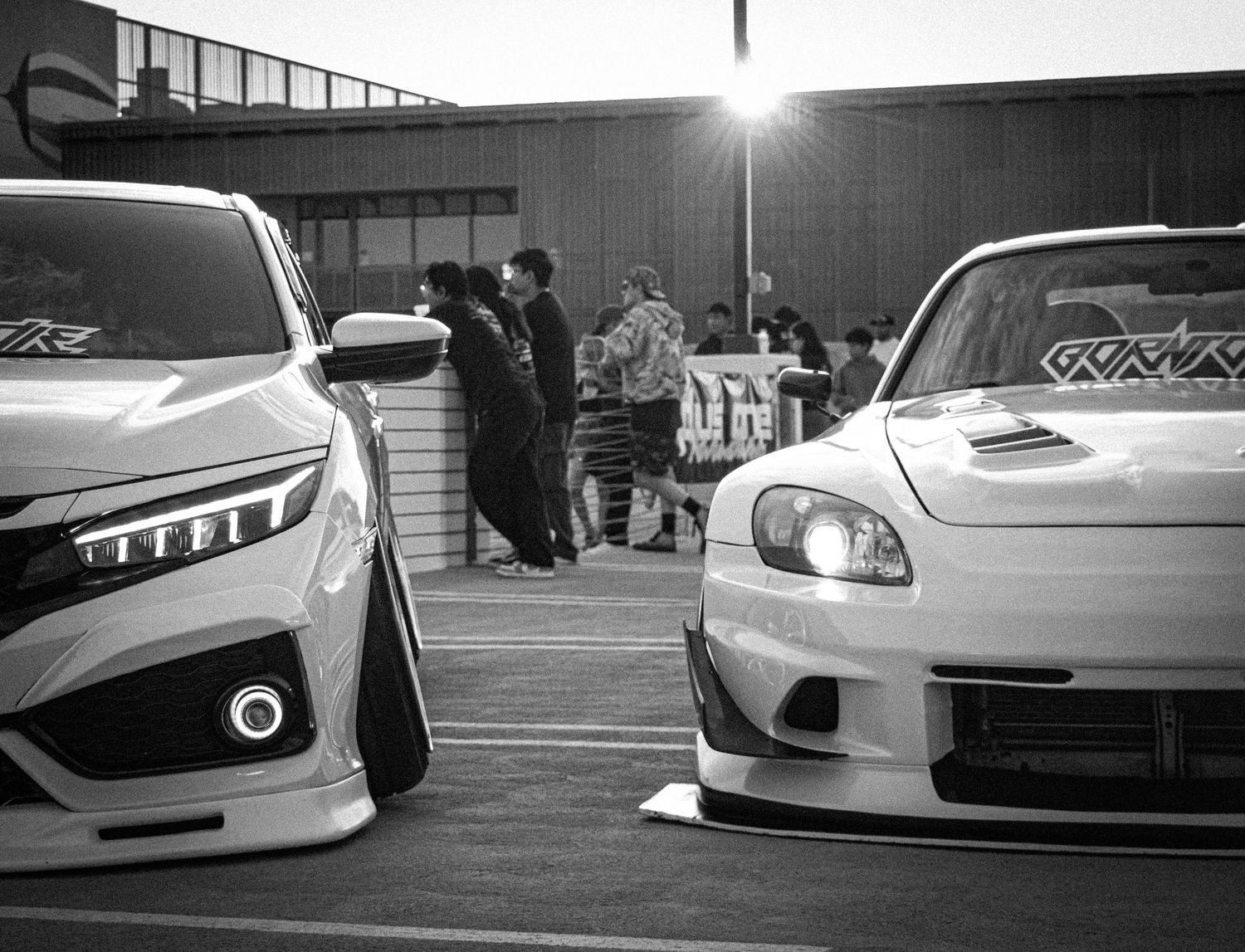 Black and white photo of two modified sport cars parked at a car meet with people gathered in the background.