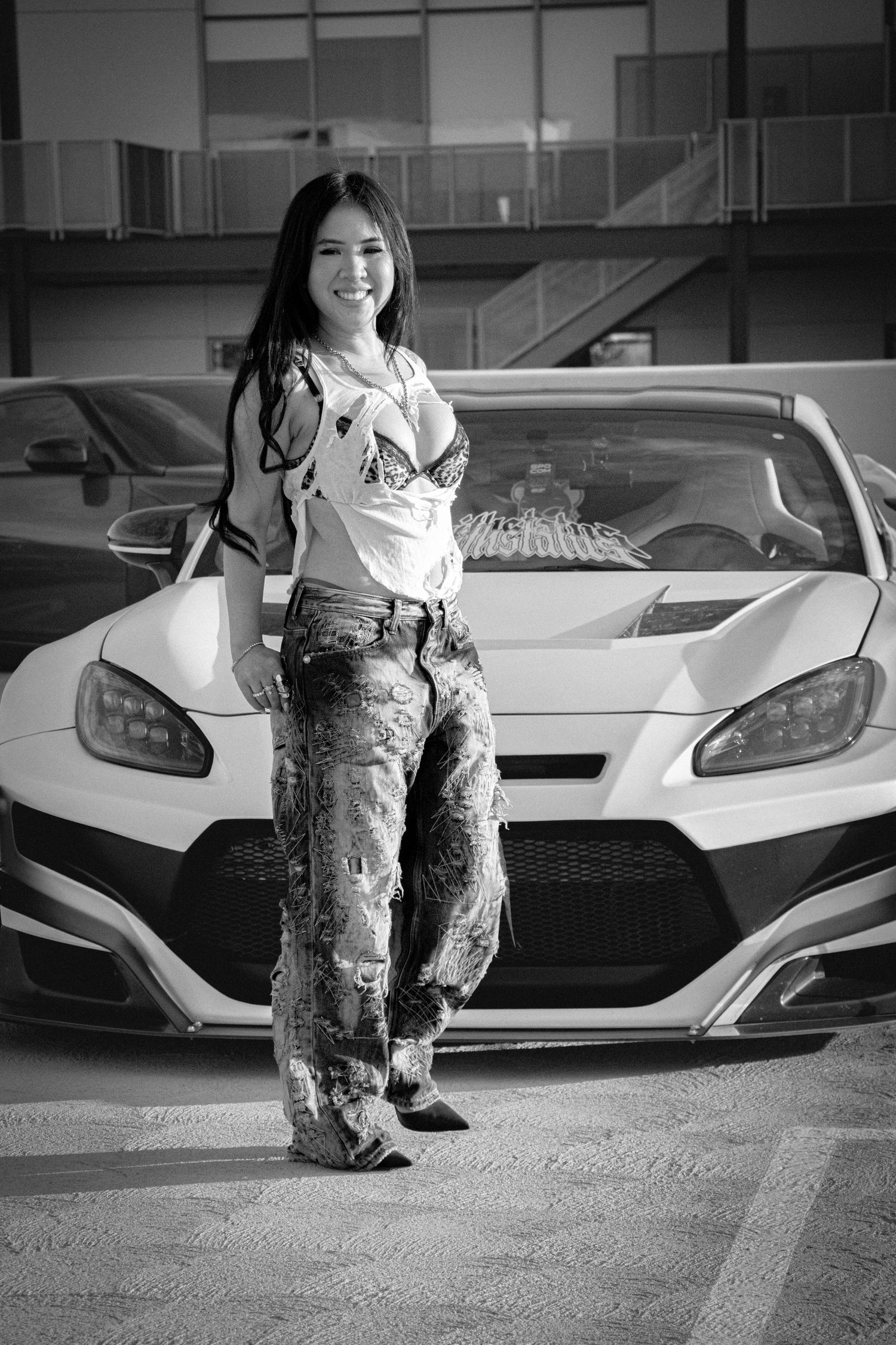 Black and white photo of a smiling young woman in patterned jeans posing in front of a white sports car in a parking lot.