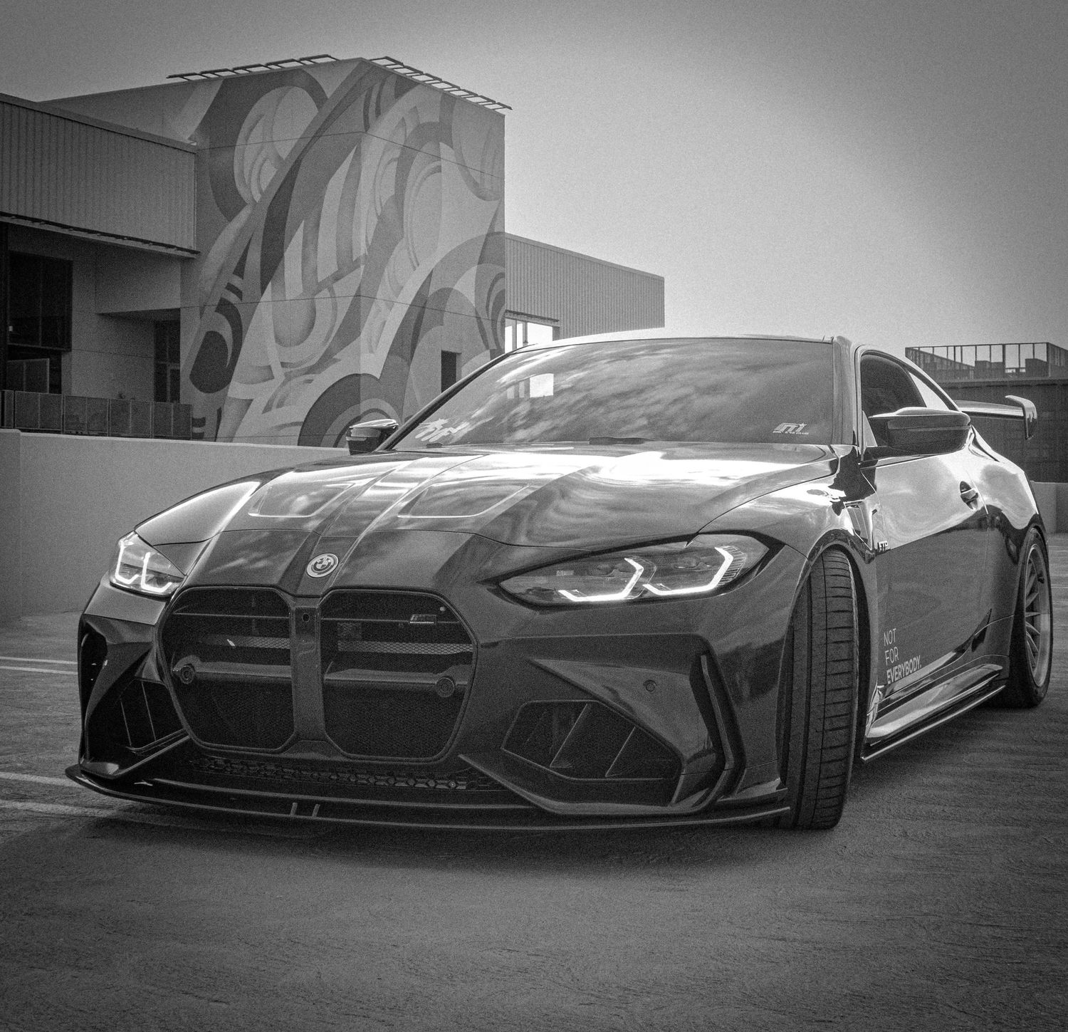 Black BMW M4 with aggressive wide body kit and custom front splitter parked in front of a mural wall.
