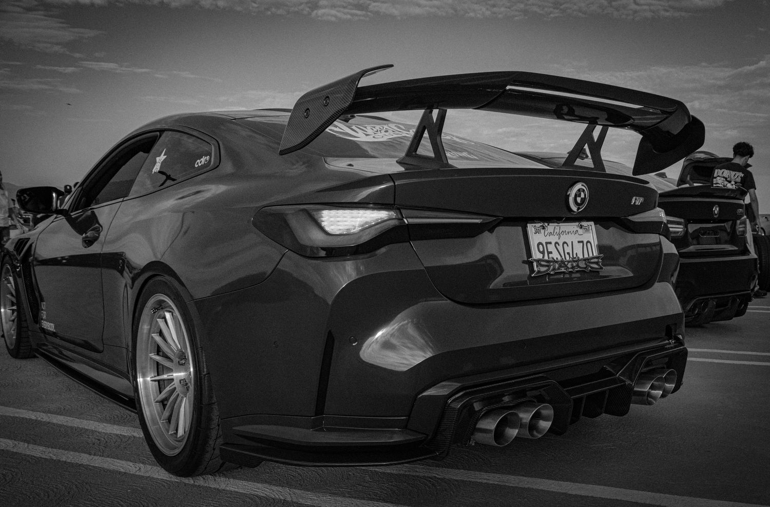 Black and white photo of a dark sports car with a large rear spoiler and quad exhaust tips in a parking lot.