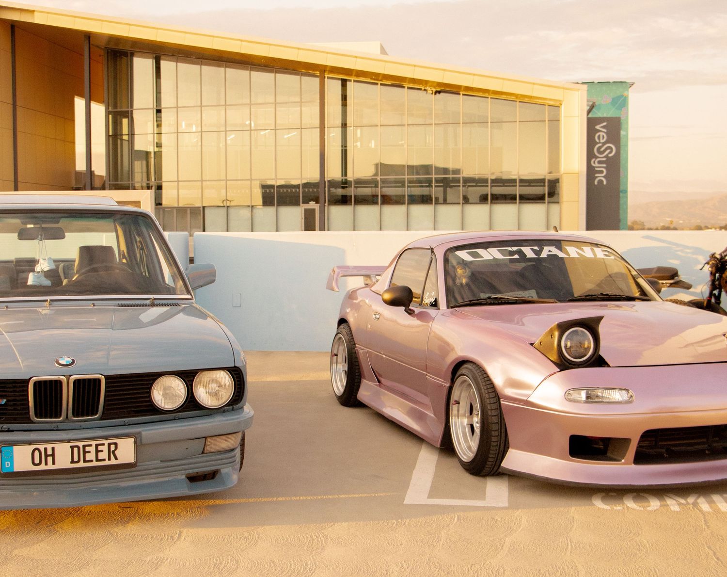 A blue BMW E28 and pink widebody Mazda MX-5 parked side by side at a car meet in a parking lot.