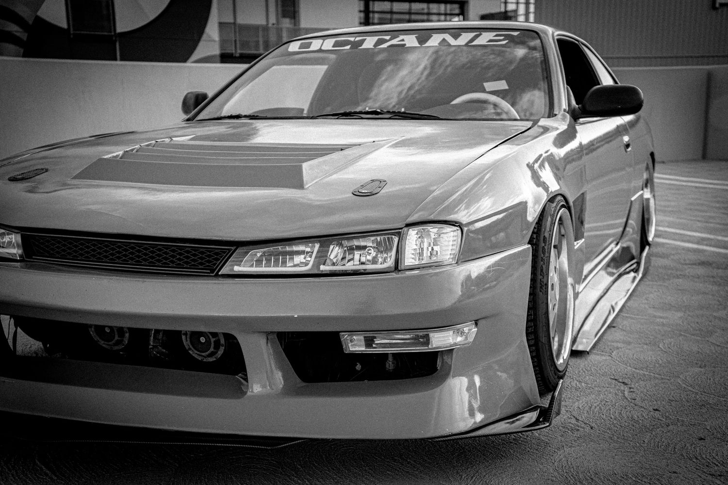Black and white photo of a modified Nissan S14 with wide body kit and custom front bumper at a car show.