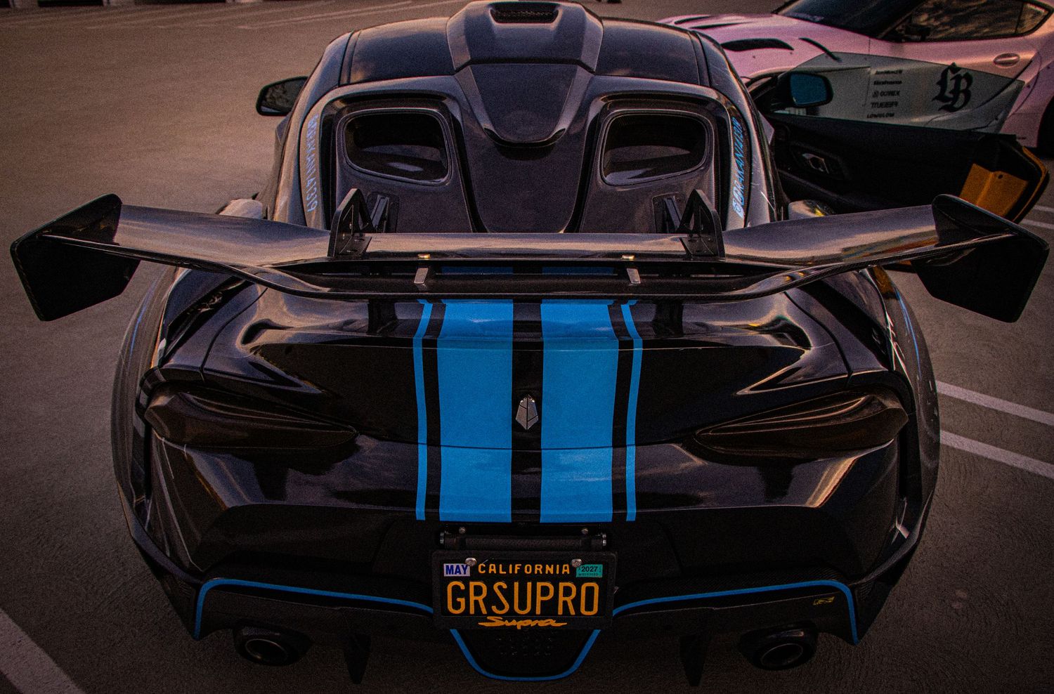 Rear view of a black and blue sports car with a California vanity plate reading GRSUPRO and large rear wing spoiler.