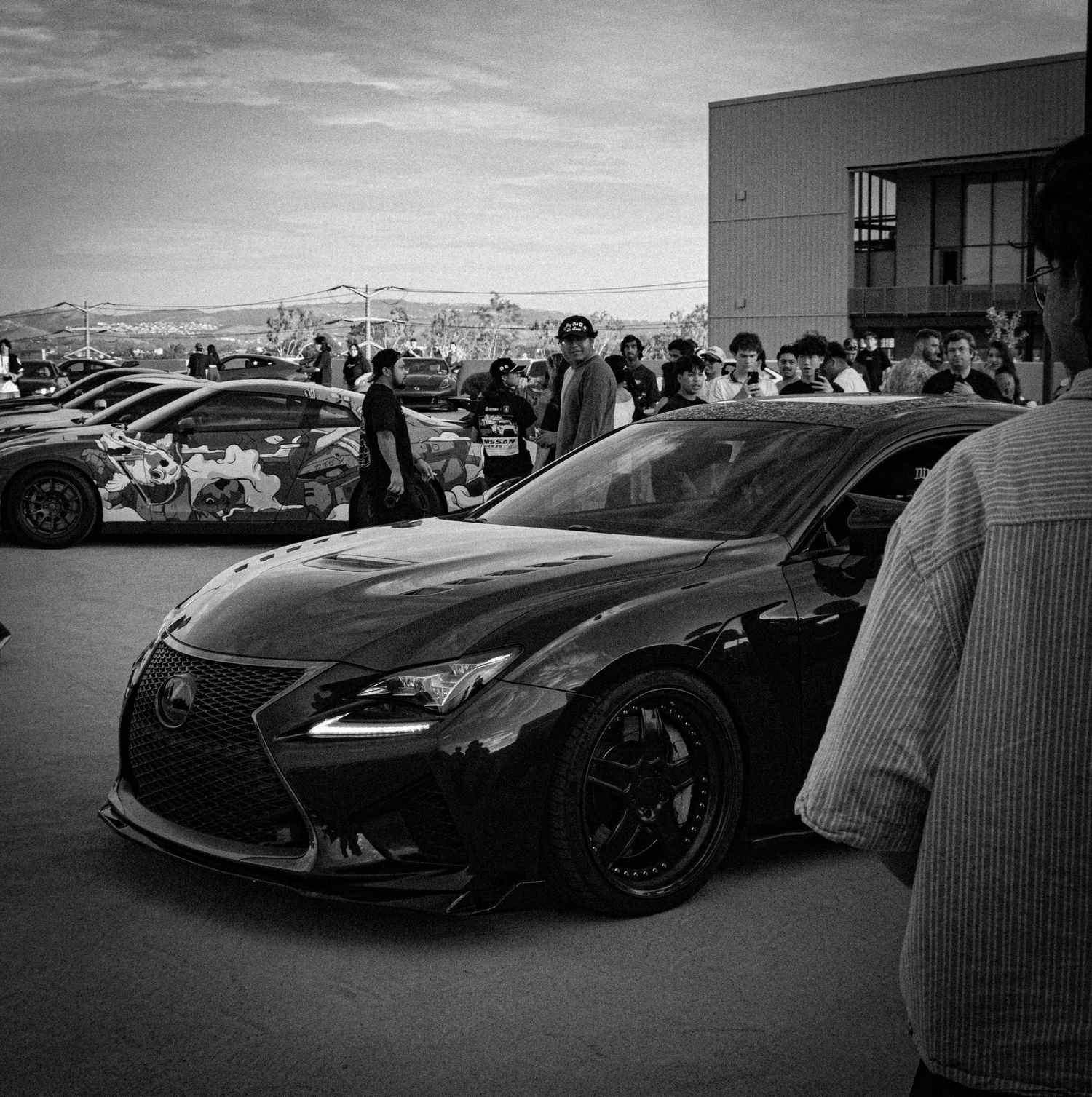 Black and white photo of a modified Lexus RC at a car meet, surrounded by spectators and custom vehicles.