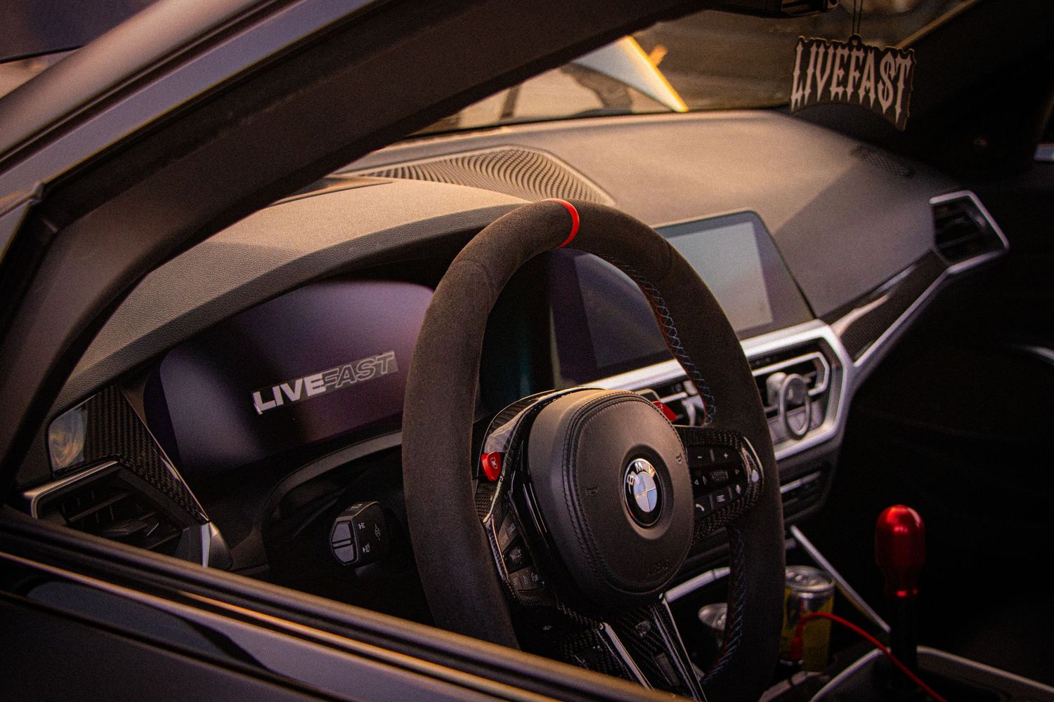 Interior view of a BMW M car featuring a carbon fiber dashboard, Alcantara steering wheel with red accents, and LiveFast branding.