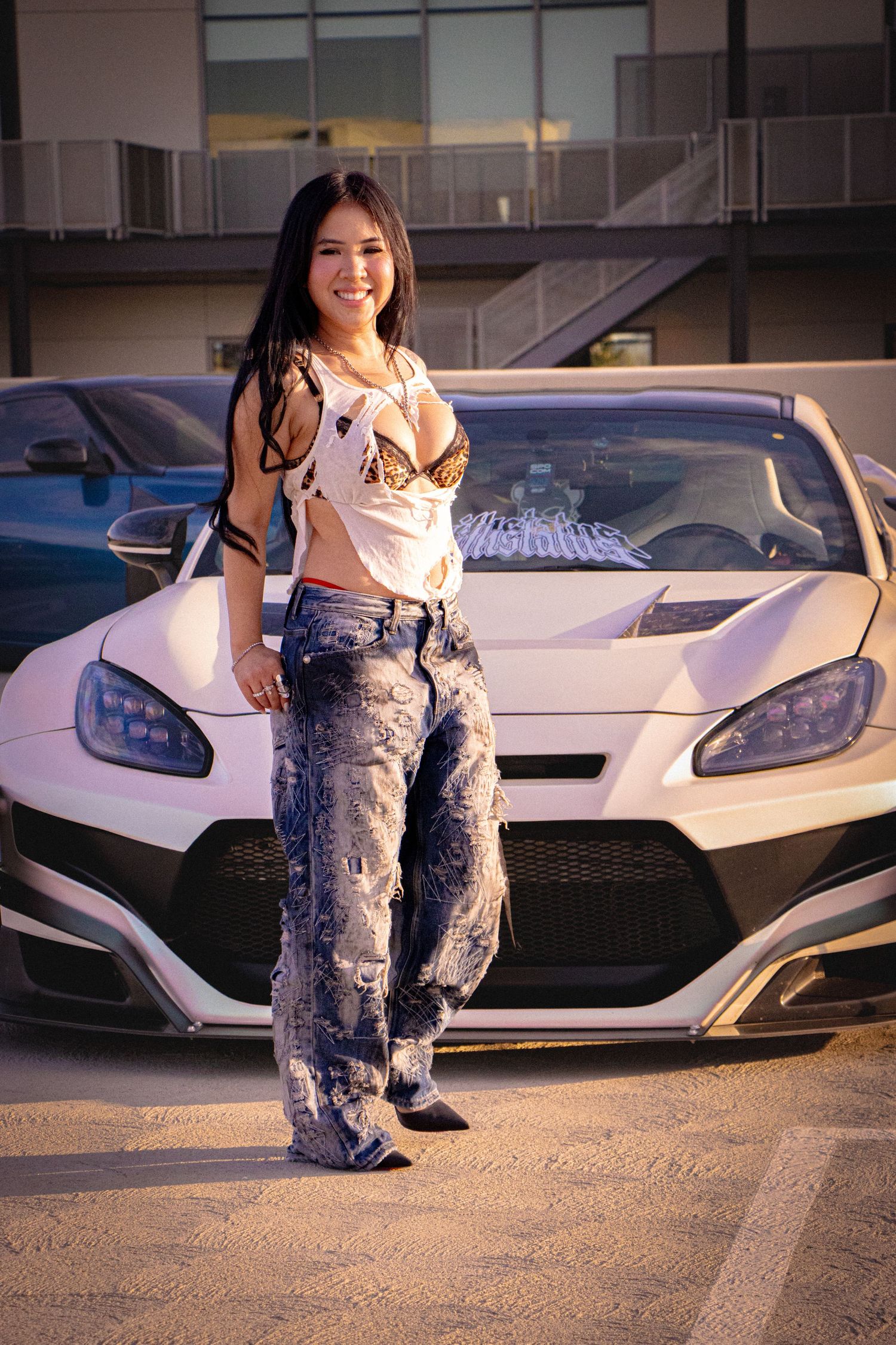 Young woman in patterned jeans and crop top posing confidently in front of a white sports car in a parking lot.