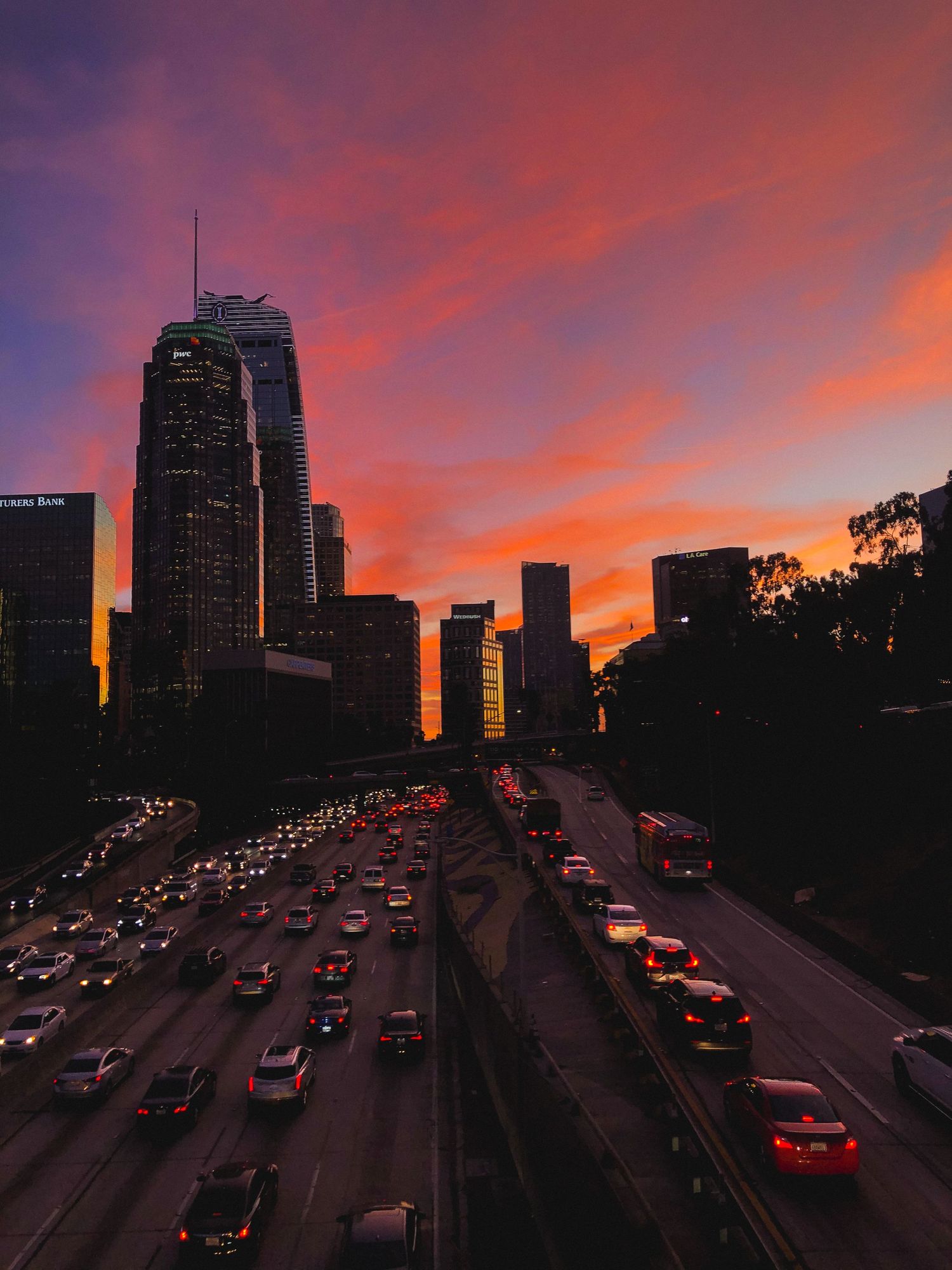 Los Angeles skyline silhouetted against dramatic purple and orange sunset with heavy traffic below.