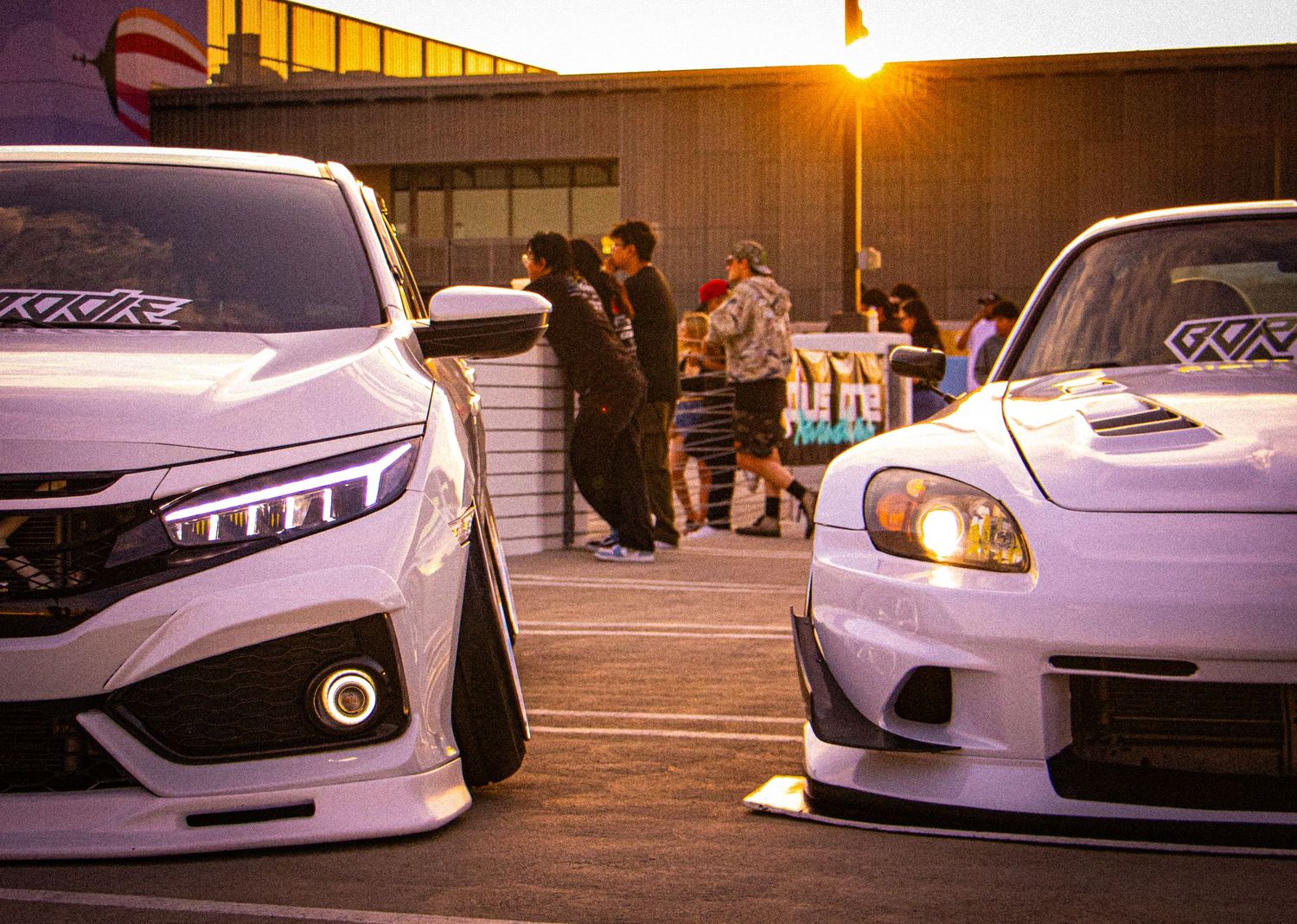 Two modified Honda cars with wide body kits parked at a car meet during sunset with people gathered in background.